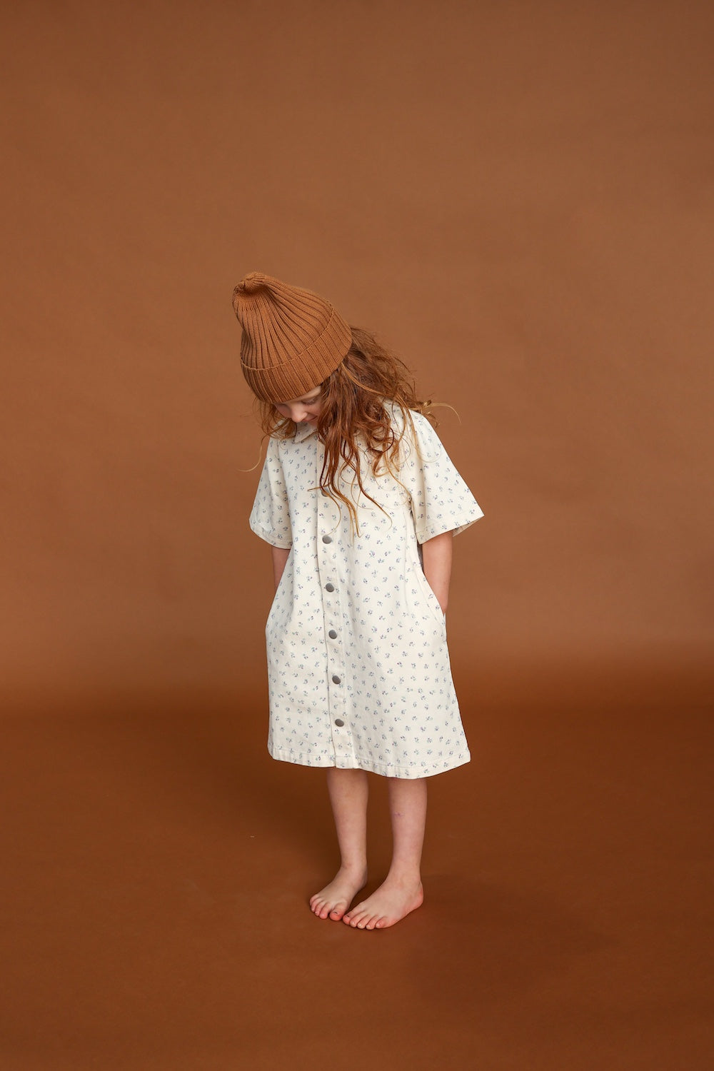 A young child stands barefoot against a brown background, wearing the GROWN - Collar Dress Garden by GROWN, a 100% cotton white dress with small floral prints and a collar detail, paired with a brown knit beanie and hands in pockets.
