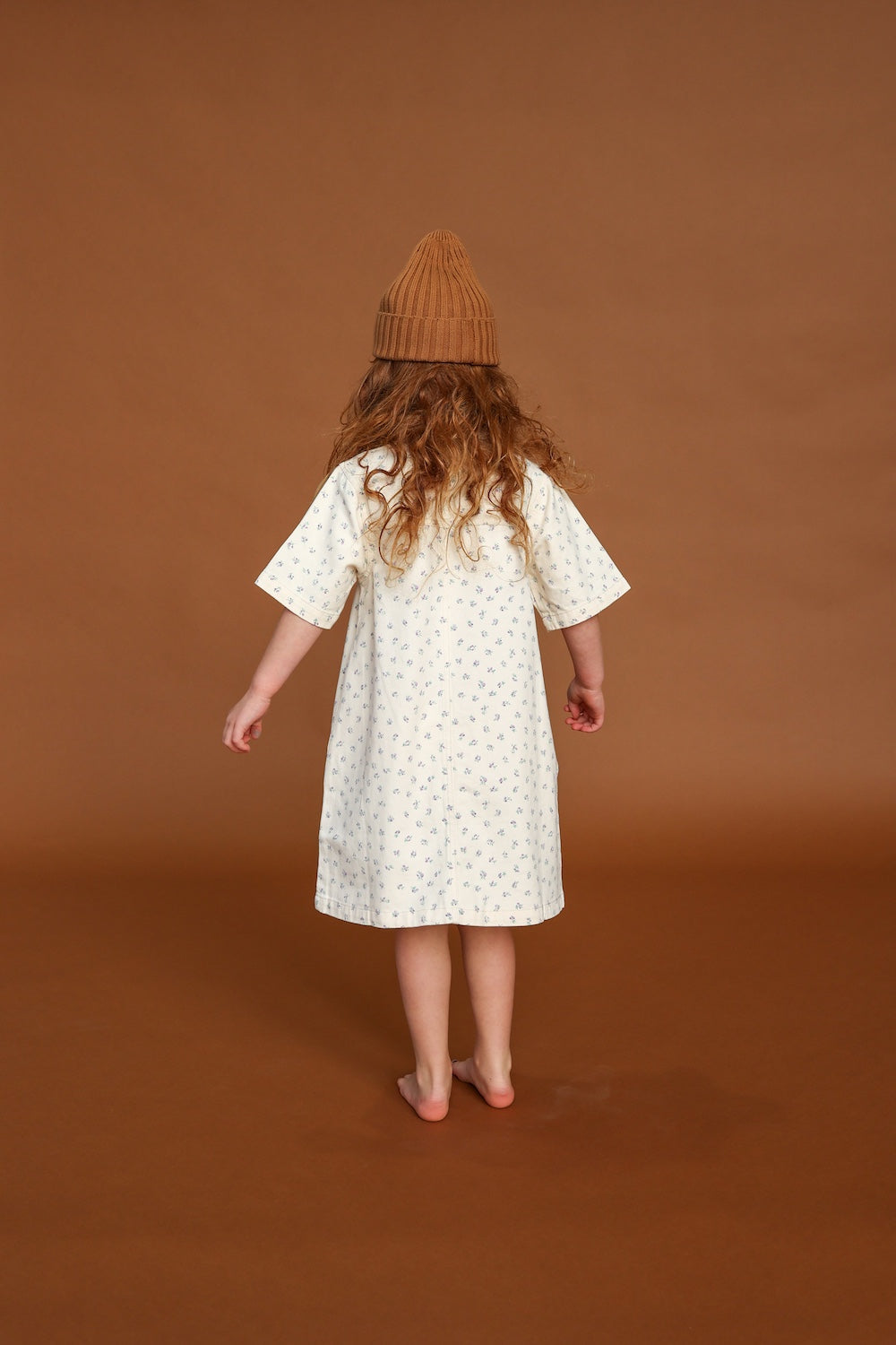 A young child with long curly hair, wearing the GROWN Collar Dress Garden and a brown knit hat, stands barefoot facing away on a brown background.