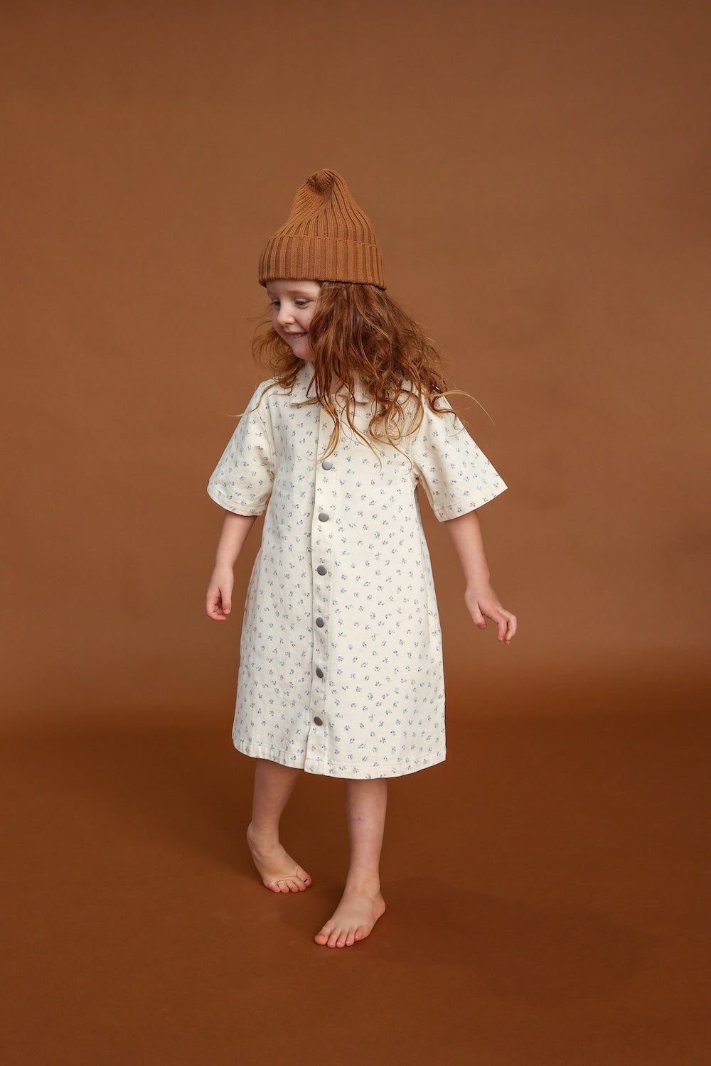 A young girl with long wavy hair wears the GROWN Collar Dress Garden—a white 100% cotton, floral-patterned dress with collar detail and press snaps—paired with a tan knit beanie, standing barefoot against a brown background and smiling.