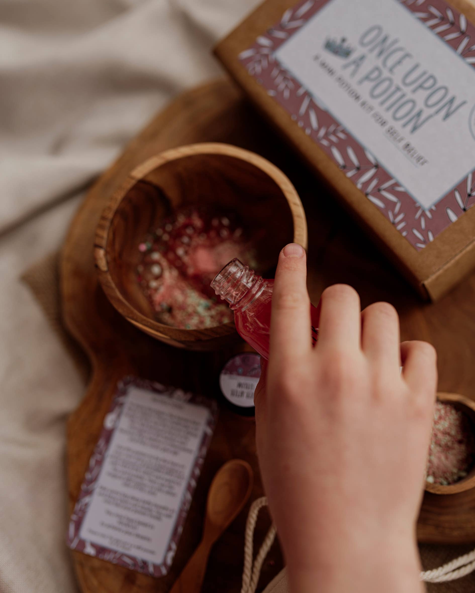 A hand pours a small bottle of red liquid from The Little Potion Co's MINI Once Upon A Potion Kit into a wooden bowl of powder, perfect for imaginative play. A box labeled "Once Upon A Potion" and a card await on a wooden board, ready to whisk you to enchanted realms.