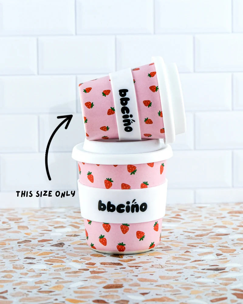 Two BBcino ~ Reusable Babycino Cup 120ml Berry BB cups with strawberry patterns and white lids rest on a tiled surface. Crafted from sustainable bamboo, one is stacked on the other, featuring an arrow and "THIS SIZE ONLY." The backdrop showcases classic white subway tiles.