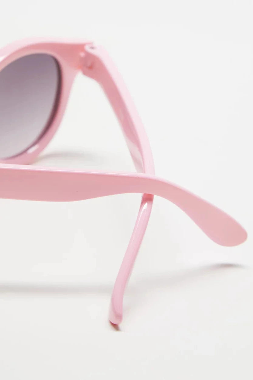 Close-up of the Baby Pixie Sunglasses Pink Stripe by FRANKIE RAY, featuring round, impact-resistant lenses with a light pink rubber frame. The slightly curved arms of these sunglasses offer 100% ultraviolet protection and are positioned on a white surface.