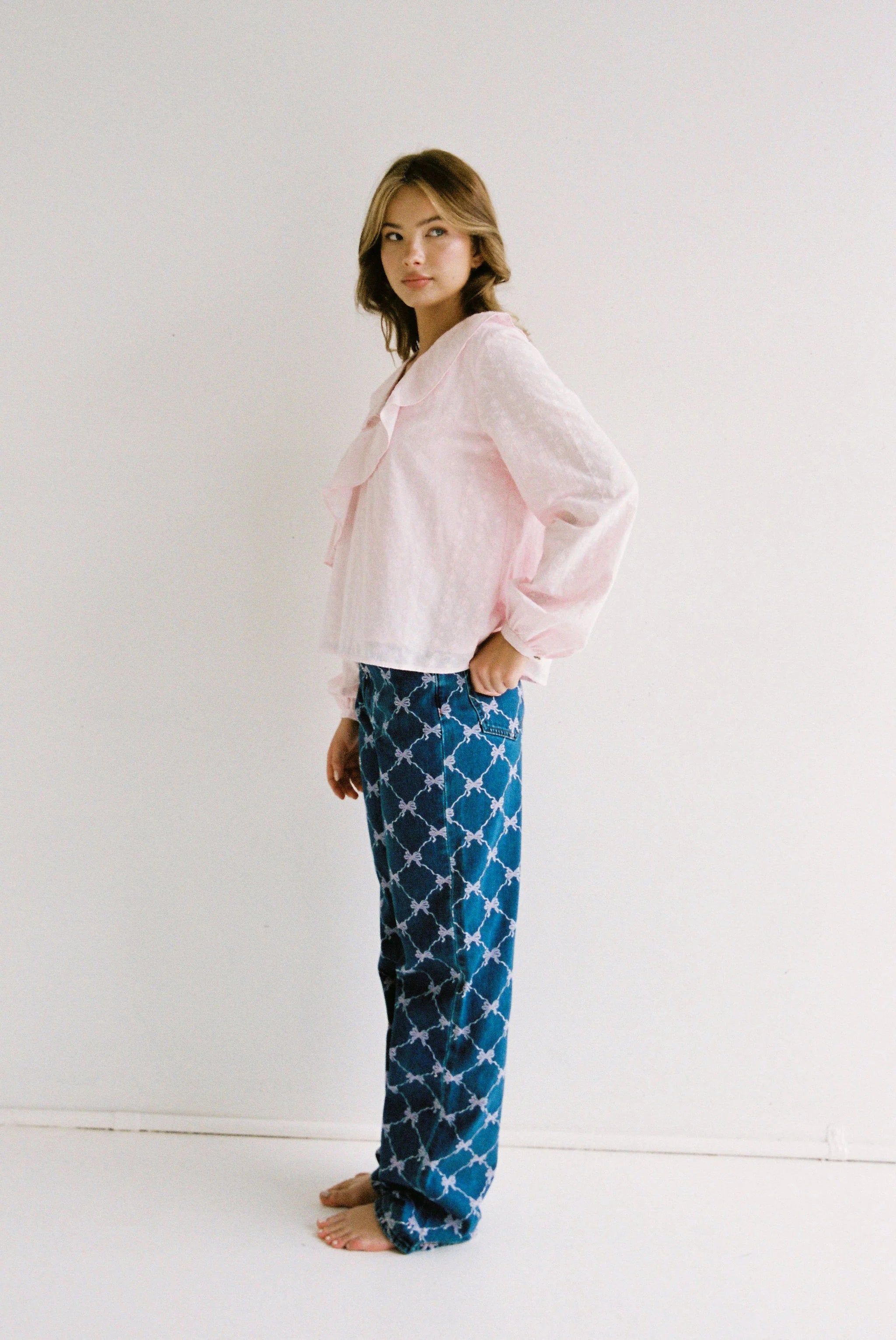 A person stands barefoot against a white background, wearing a light pink ruffled blouse and the JUNI - Sabrina Jean Bow Print (Regular Length) pants by JUNI. They face slightly to the side and look toward the camera.