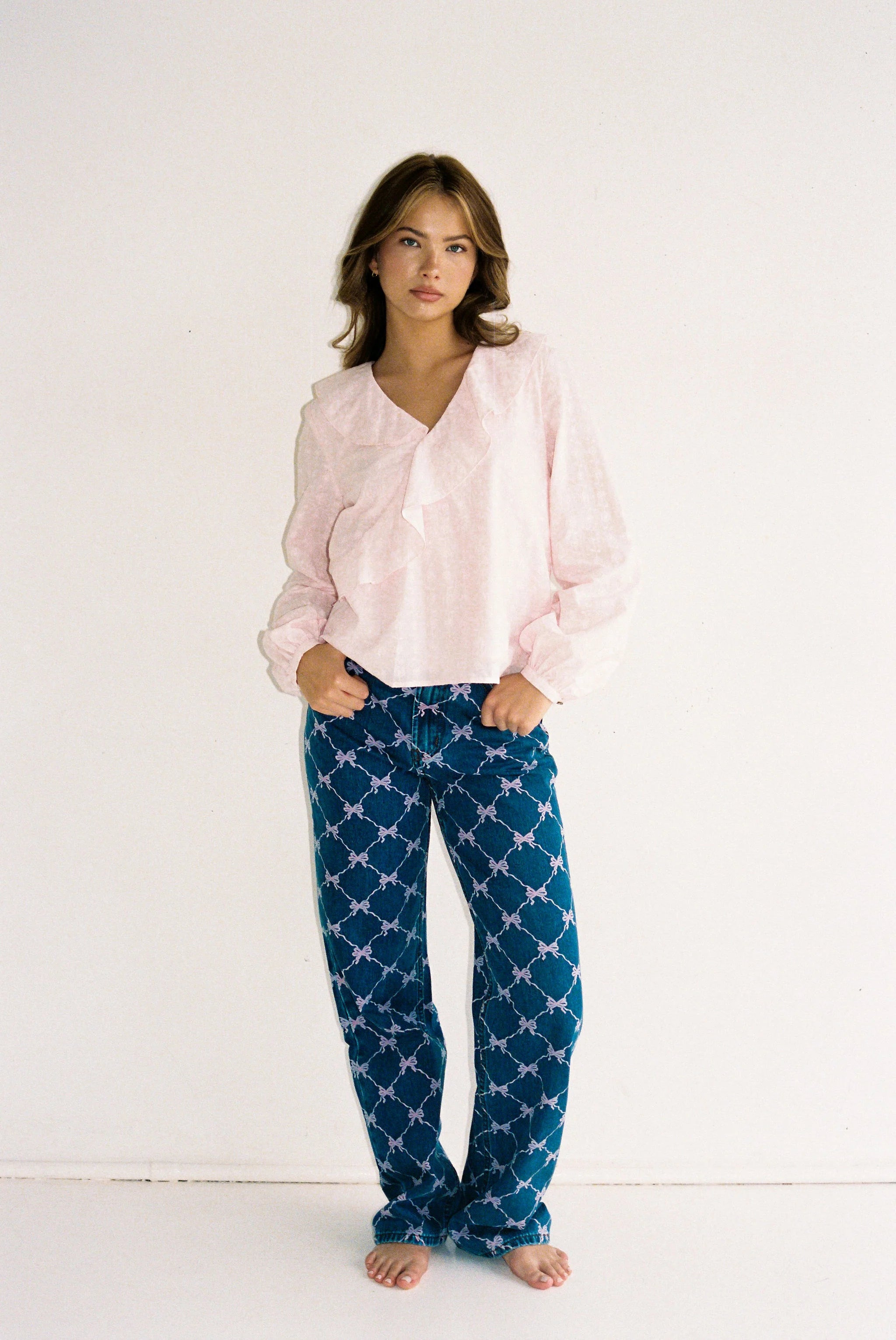 A young woman with loose, wavy hair and a neutral expression stands barefoot against a plain white wall, wearing a light pink long-sleeve blouse and JUNI's Sabrina Jean Bow Print (Regular Length) by JUNI.