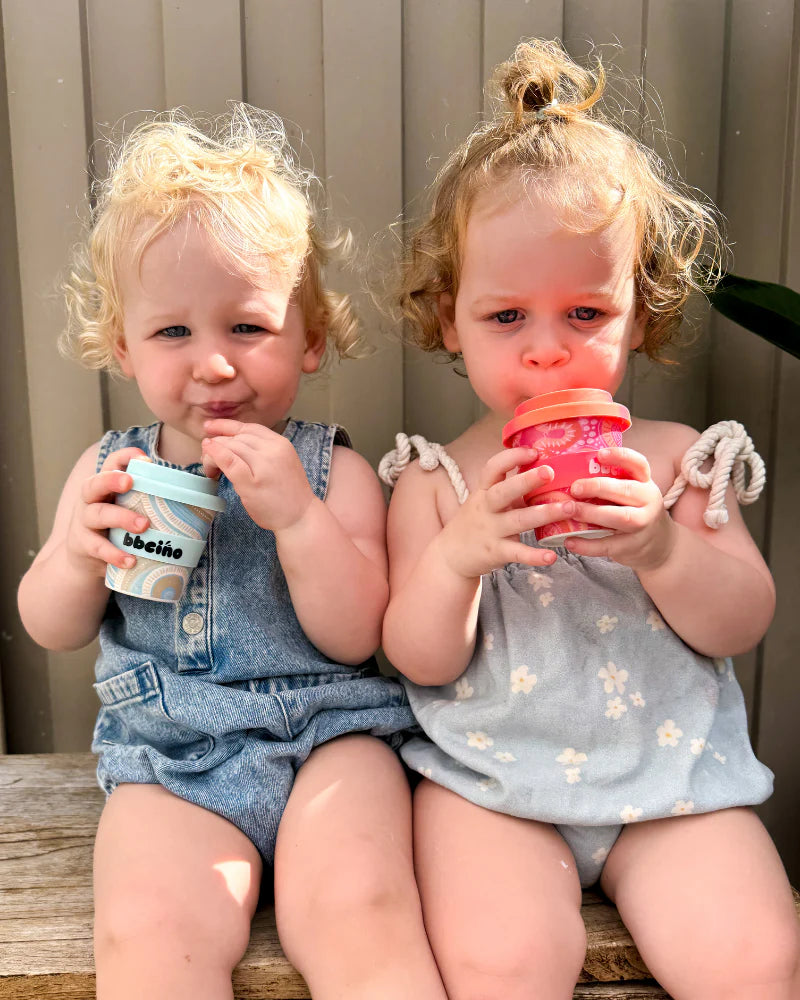 Two young children with curly blonde hair sit together on a bench, each holding a colorful BBcino - Reusable Babycino Cup 120ml Coast by BBCINO, dressed in light summer outfits and looking at the camera with calm expressions.