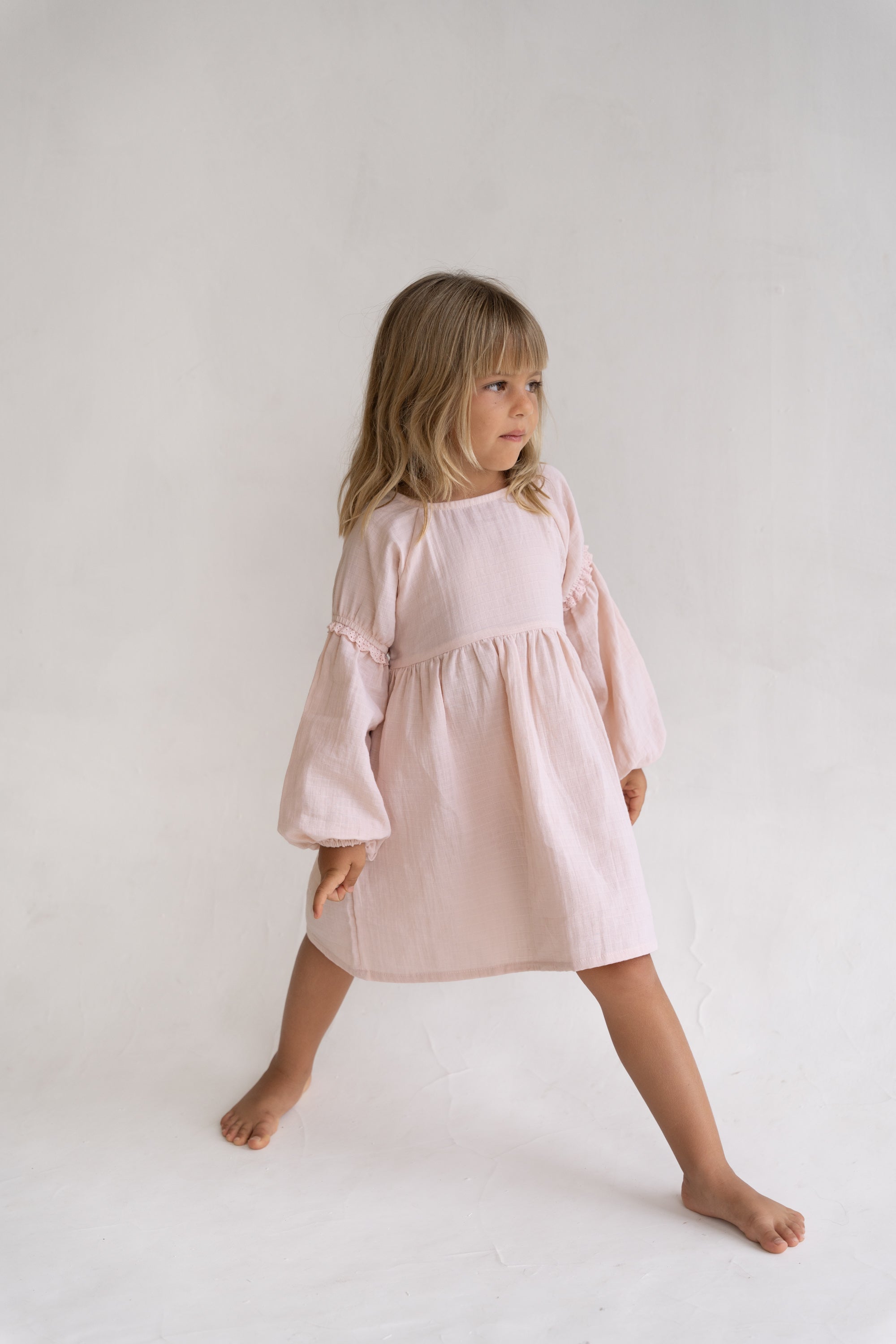 A young girl with light brown hair stands barefoot against a white backdrop, looking to the side and wearing the Illoura the Label - Maggie Dress Pink Salt by ILLOURA THE LABEL, featuring long sleeves and a soft pink hue.