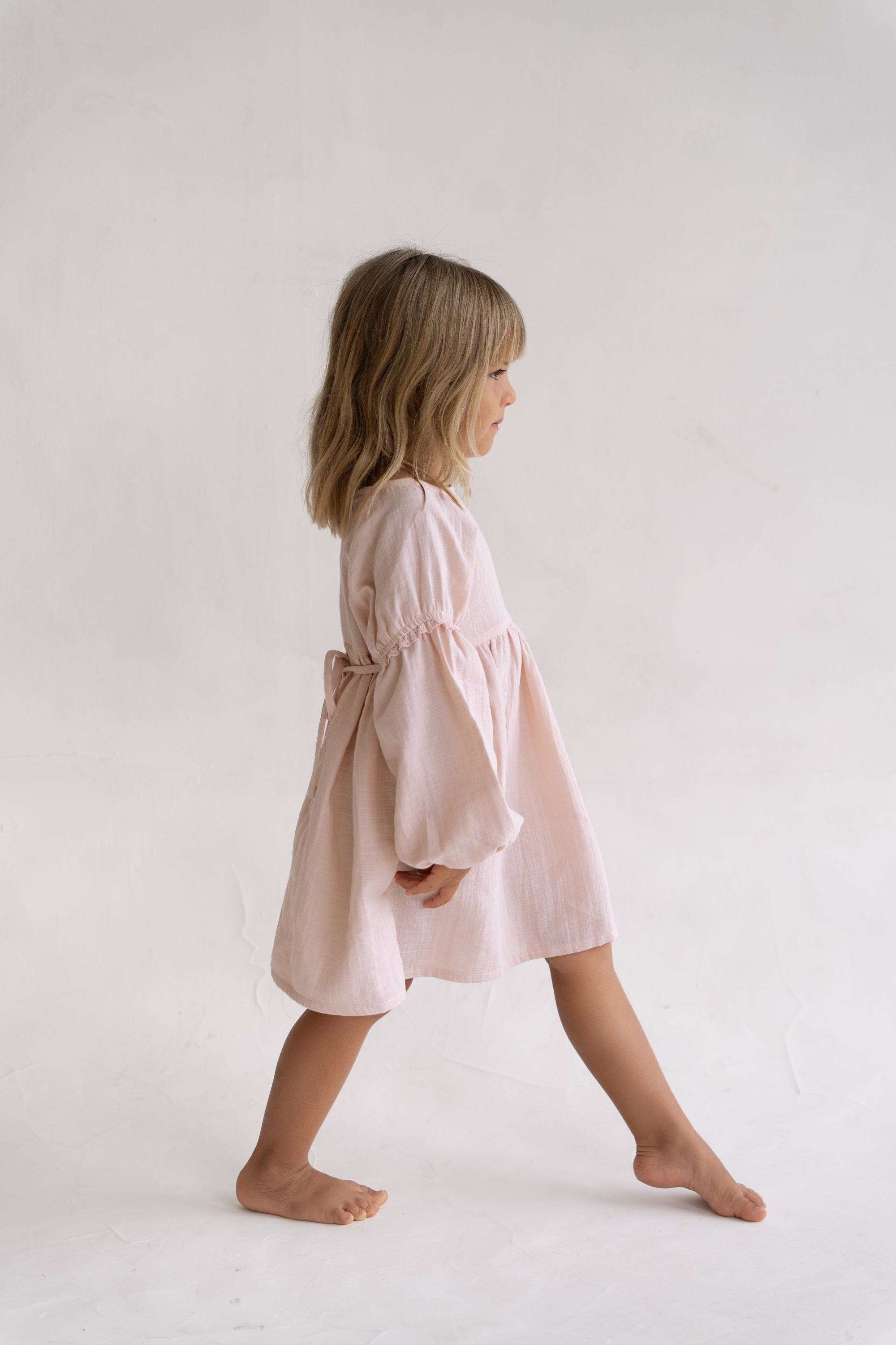 A young girl with blonde hair walks barefoot in profile against a plain white background, wearing the Illoura the Label - Maggie Dress Pink Salt by ILLOURA THE LABEL.