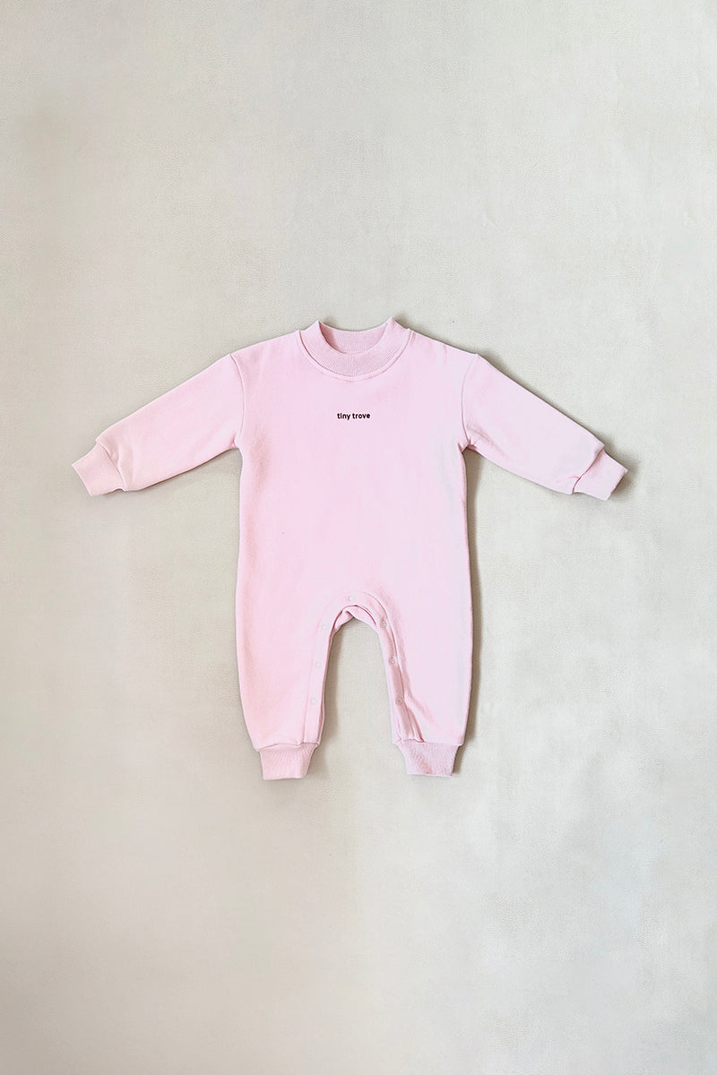 The TINY TROVE Otto Puff Circle Logo Romper Pink is a light pink, long-sleeve onesie with ribbed cuffs and a puff logo on the chest, shown laid flat on a neutral background.