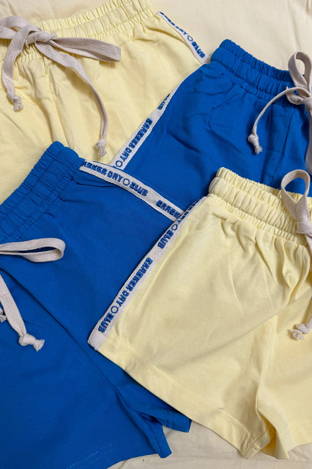 Four pairs of CRACKER DAY CLUB cotton kids shorts are displayed: two yellow, one solid blue, and one Racer Club Shorts Sunshine style in blue with white trim and side text. All have elastic waists and drawstrings.