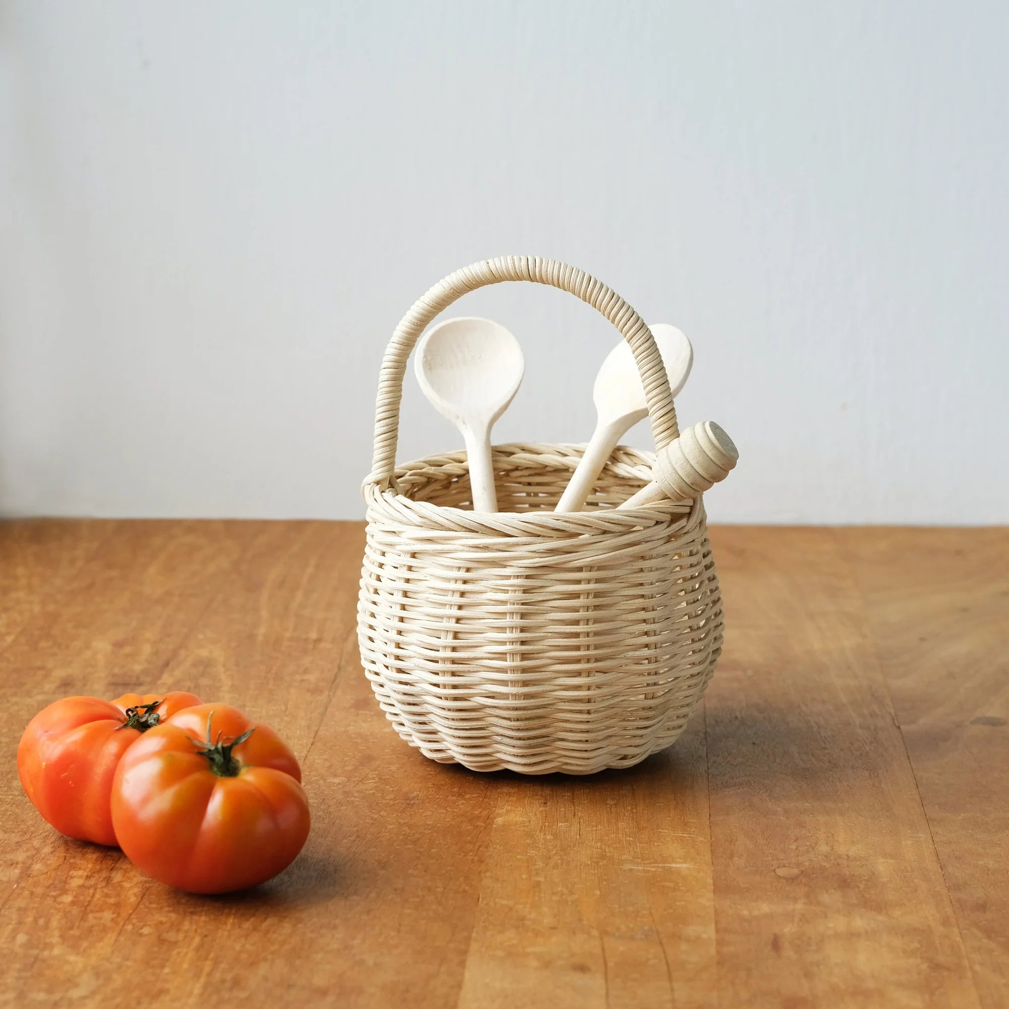 The Olli Ella Berry Basket Straw by OLLI ELLA, crafted from natural rattan, holds three wooden utensils on a wooden surface. Two ripe tomatoes are placed beside it, set against a plain white wall.
