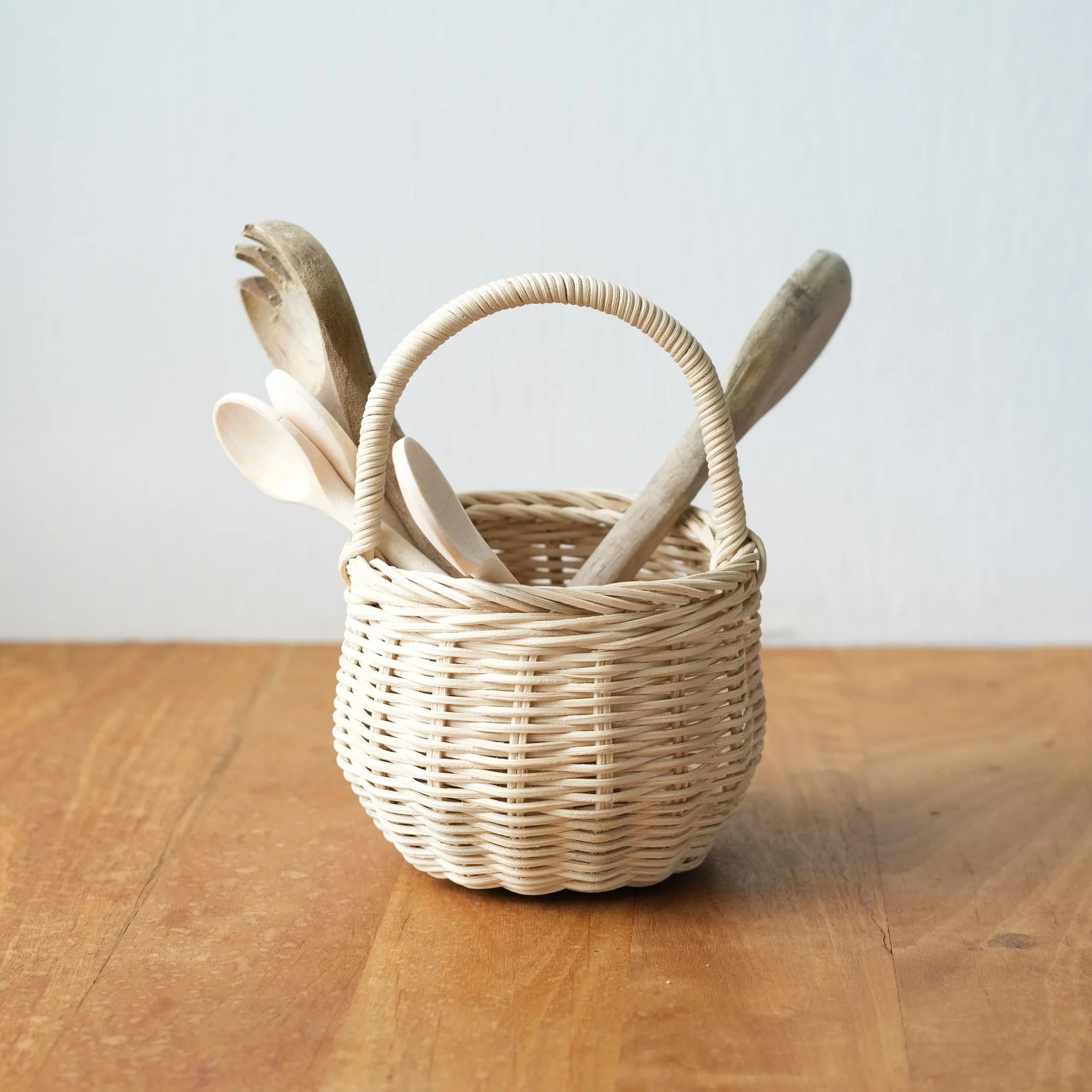 The OLLI ELLA Berry Basket Straw, a small handmade woven rattan piece with a curved handle, sits on wood holding wooden kitchen utensils like spoons and spatulas against a plain white wall.