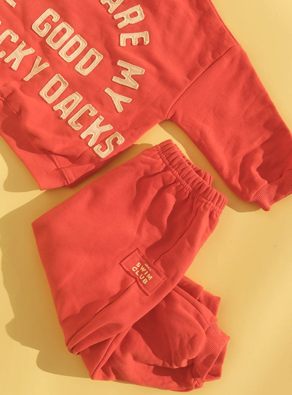 The Small Swim Club Good Tracky Dacks Red/Cream children’s set features a red sweatshirt with bold partial text and matching fleece-lined, oversized track pants with a small white patch, all displayed on a yellow surface. Brand: SMALL SWIM CLUB.