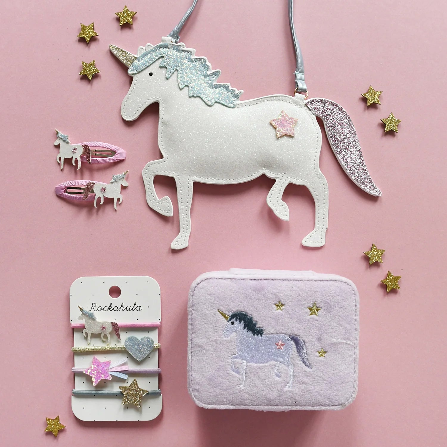 Flat lay of ROCKAHULA’s Unicorn Ponies on a pink background, featuring unicorn hair elastics, a white unicorn purse, pink hair clips, a lavender pouch, and gold glitter star decorations—perfect for kids who love glittery unicorn accessories.