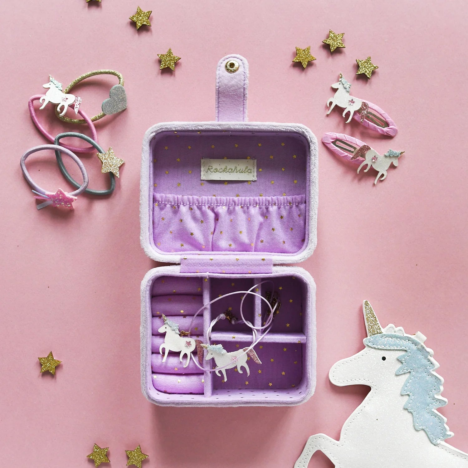 The ROCKAHULA Rockahula - Unicorn Clips are shown on a pink background, beside a pastel purple jewelry box with a unicorn necklace, star-shaped accessories, glittery unicorns, ribbon clips, and a unicorn cutout.