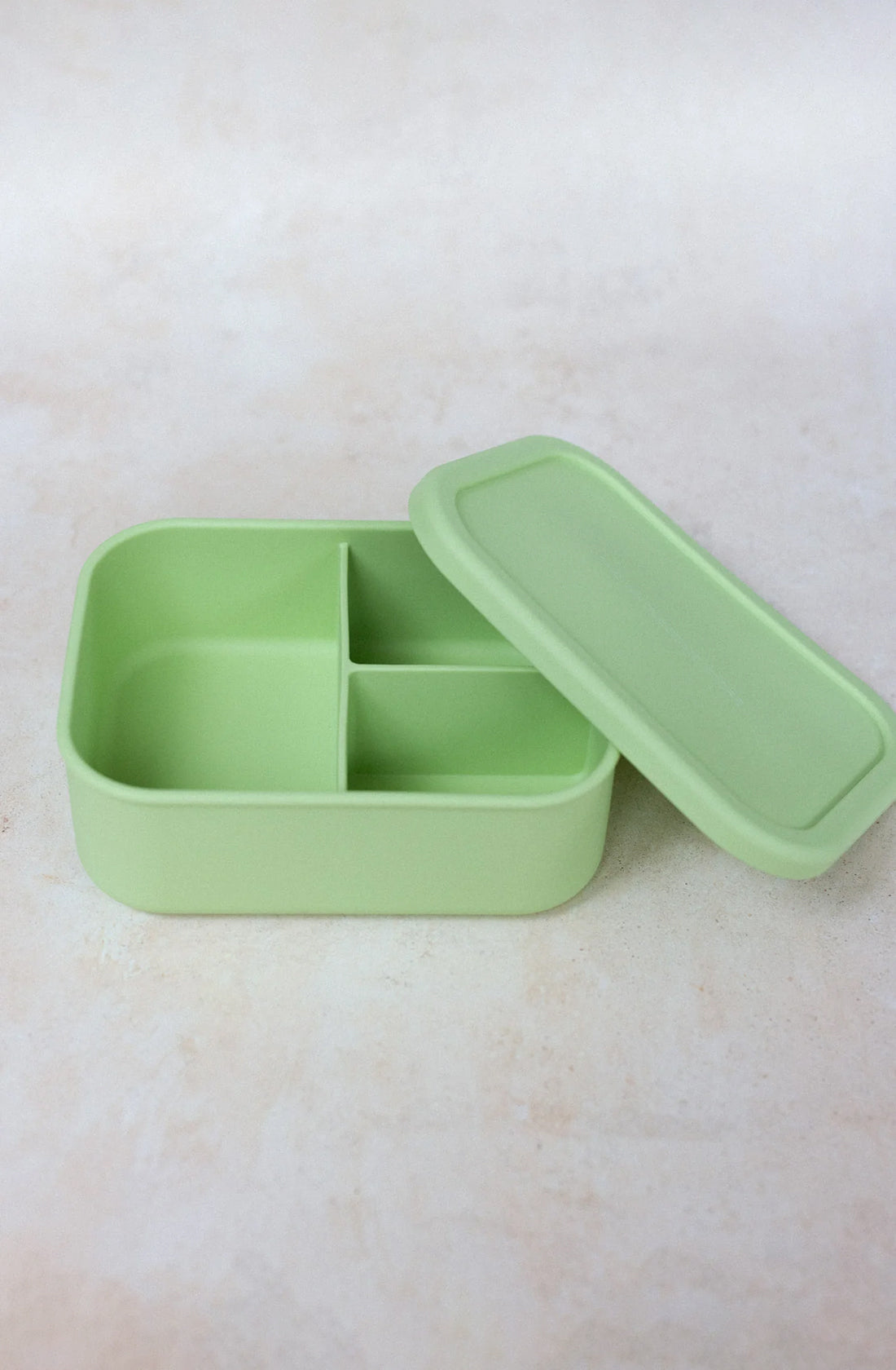A light green "Silicone Bento Lunchbox Large (1300ml)" by THE WHOLESOME STORE, featuring three airtight compartments and a matching lid, sits next to it on a light, textured surface. The lunchbox is also dishwasher safe.