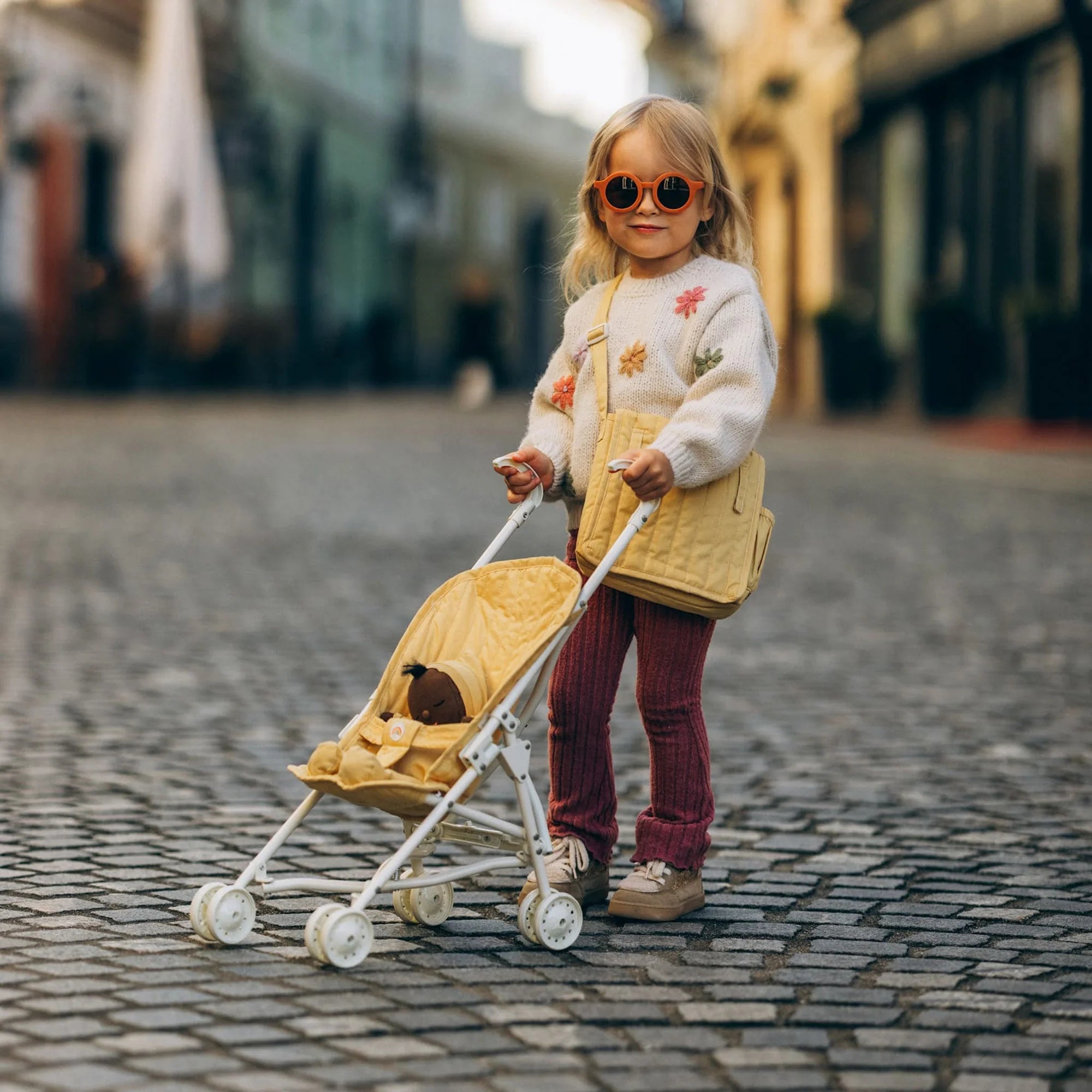 A young girl in sunglasses, a white floral sweater, and red pants stands on a cobblestone street, pushing the OLLI ELLA Sollie Stroller Honey with a small stuffed animal inside.