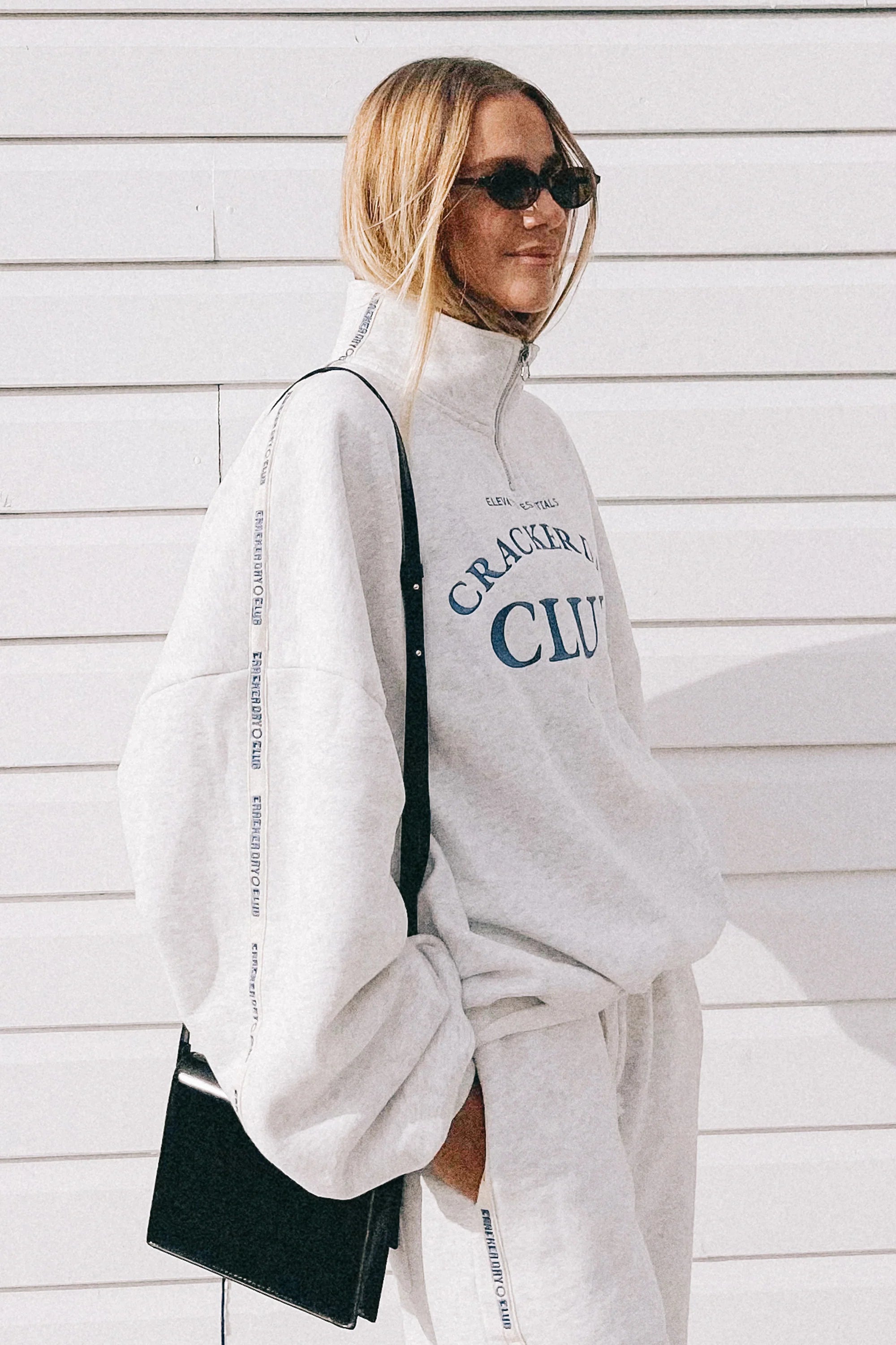 A woman in an oversized CRACKER DAY CLUB Cloud Quarter Zip Sweater in Grey Marle stands before a white wall. She wears matching sweatpants, has long blonde hair, sunglasses, and carries a black shoulder bag.
