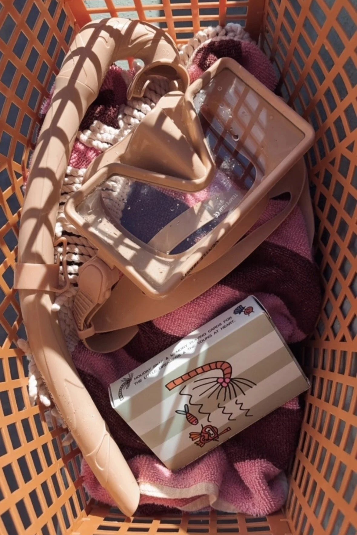 A brown plastic basket holds a set of LULU PAWLIK’s Palm & Coco On Vacay Kids Playing Cards, pink and beige hangers, a snorkel mask, and a folded purple towel. Sunlight casts playful shadows across the items.