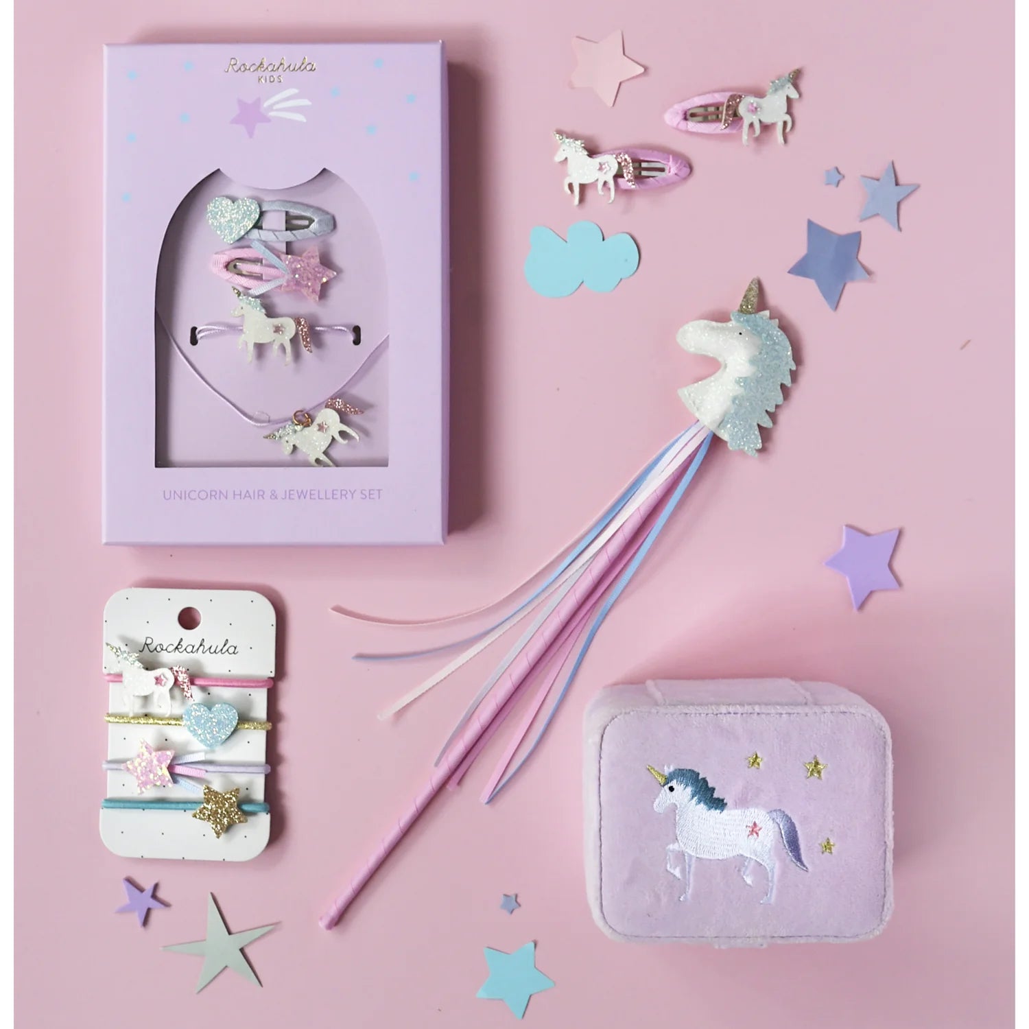 A pastel-themed flat lay features ROCKAHULA's Rockahula - Unicorn Ponies hair elastics, glittery unicorn hair clips, a pink-boxed jewelry set, a unicorn wand, and a unicorn-patterned box amid star and heart confetti on pink.