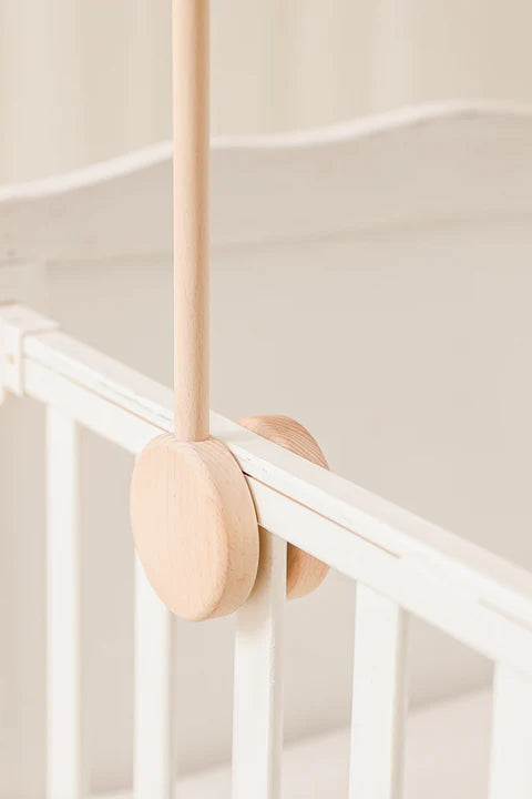 Close-up of the ROOMI & ME Roomi & Me - Wooden Mobile Stand clamped to a white crib rail, securing a vertical rod. This wooden mobile stand mounts easily on cots. Softly blurred background.