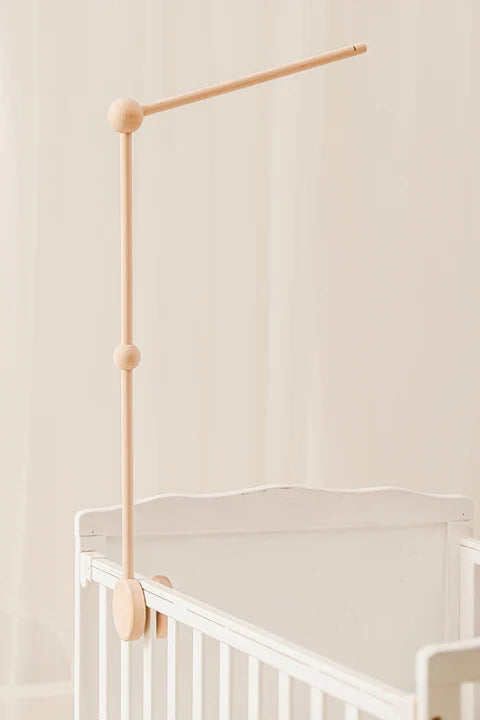 The ROOMI & ME Roomi & Me - Wooden Mobile Stand is mounted upright on the side of a white baby crib, its horizontal arm extending over the crib, suitable for cots, with a light beige background.