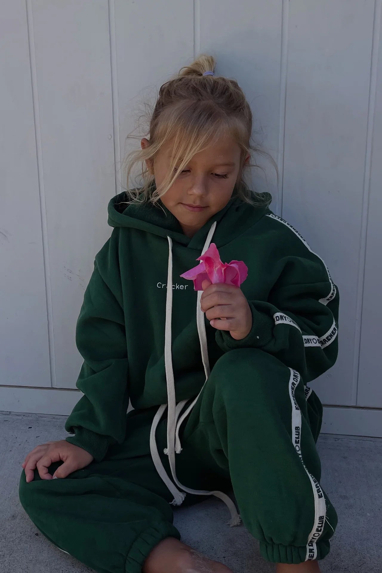A young girl with blonde hair wears CRACKER DAY CLUB's Kids Cloud Racer Sweatpants in Forest as she sits against a light wall, holding and thoughtfully looking at a pink flower.