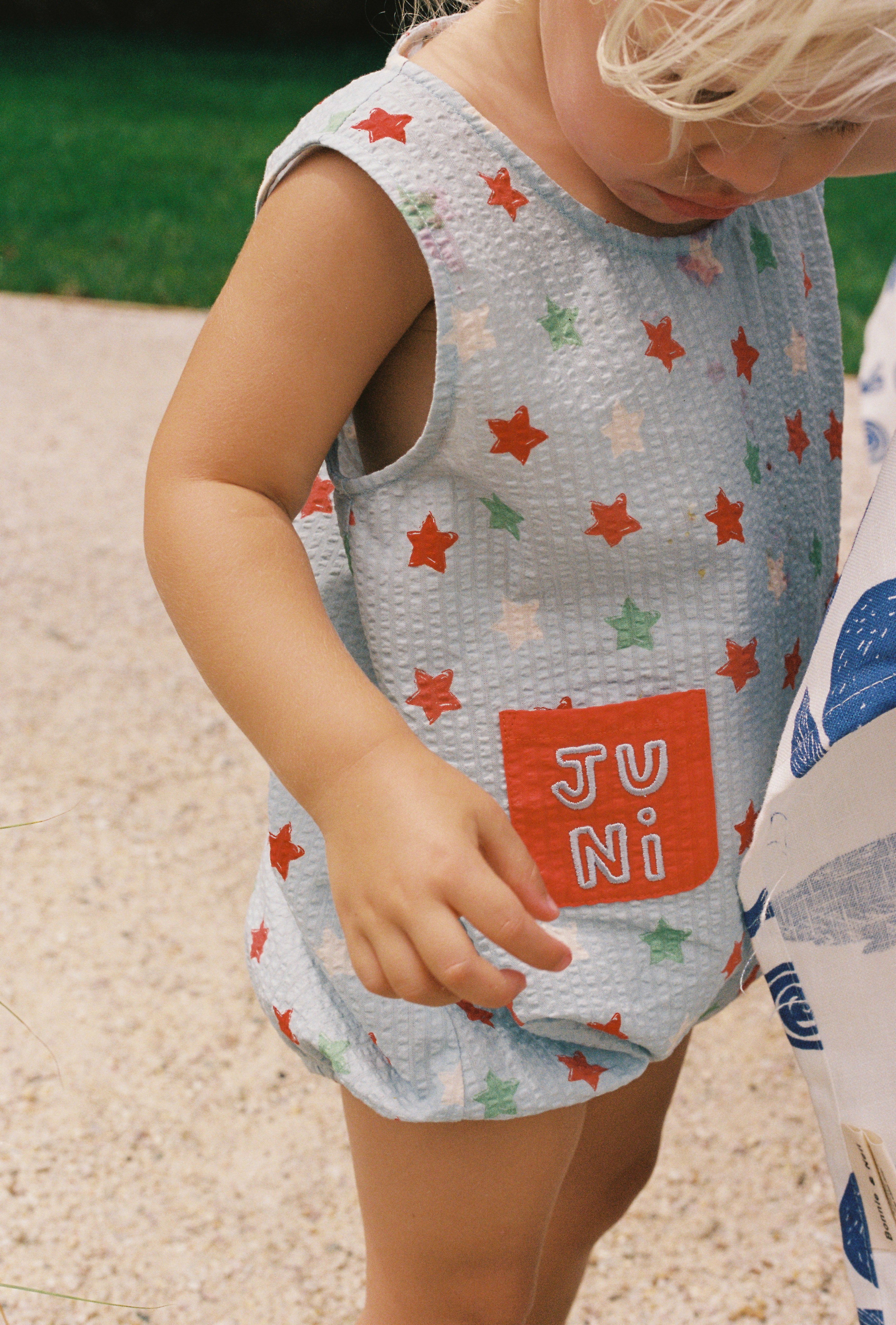 Standing outside, a toddler sports the Levi Romper from JUNI JNR. This sleeveless outfit showcases red, green, and blue stars on its seersucker fabric, complete with a practical orange pocket displaying the "JUNI" brand name. The child's light blonde hair is gently tousled against a background of grass and concrete.