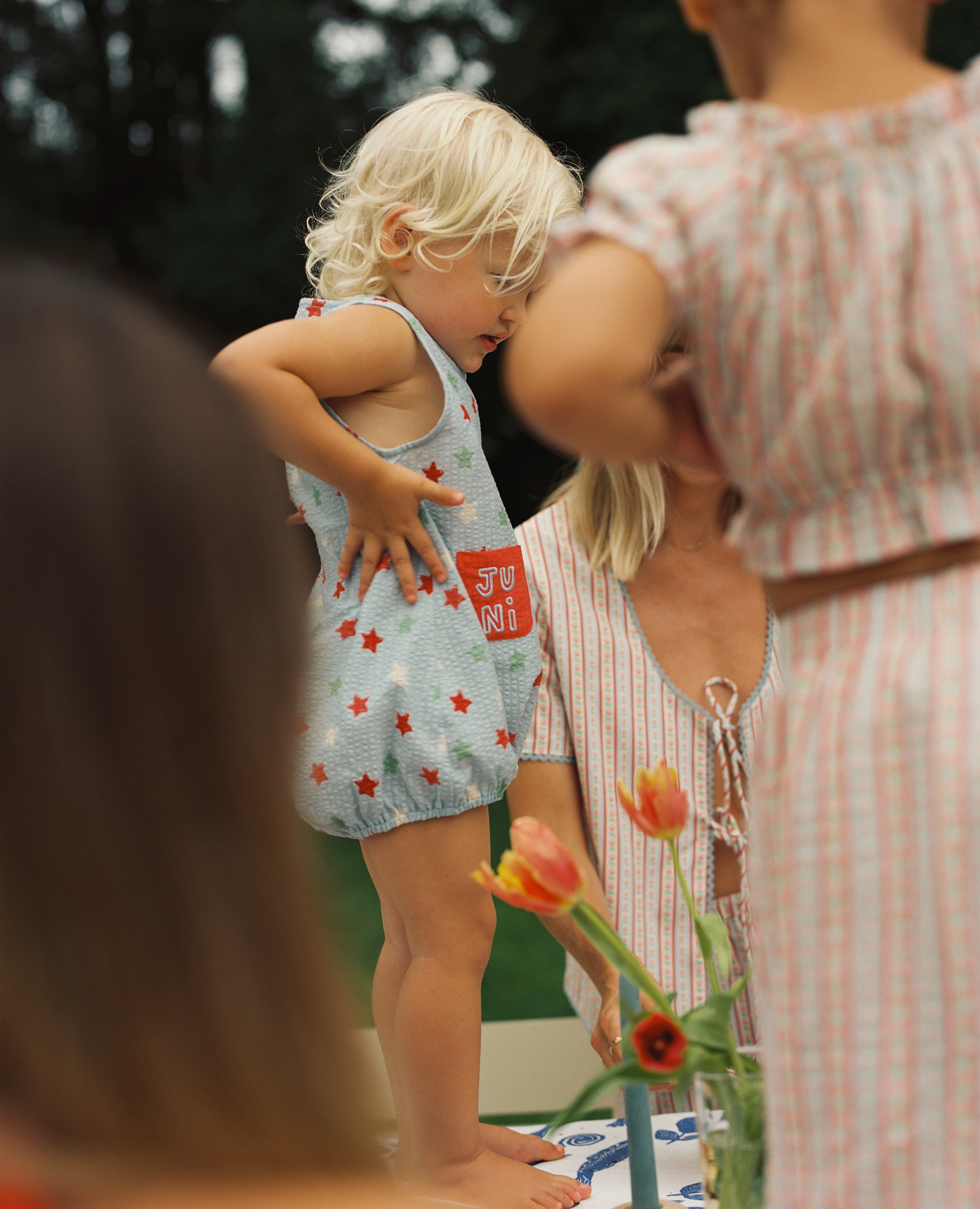 A young child with blond hair stands on a chair at a table outdoors, wearing the adorable Levi Romper by JUNI JNR, an outfit crafted from ethically made materials featuring a cute star pattern. Two adults are partially seen nearby, and tulips in a vase add charm to the scene, with blurred greenery in the background.