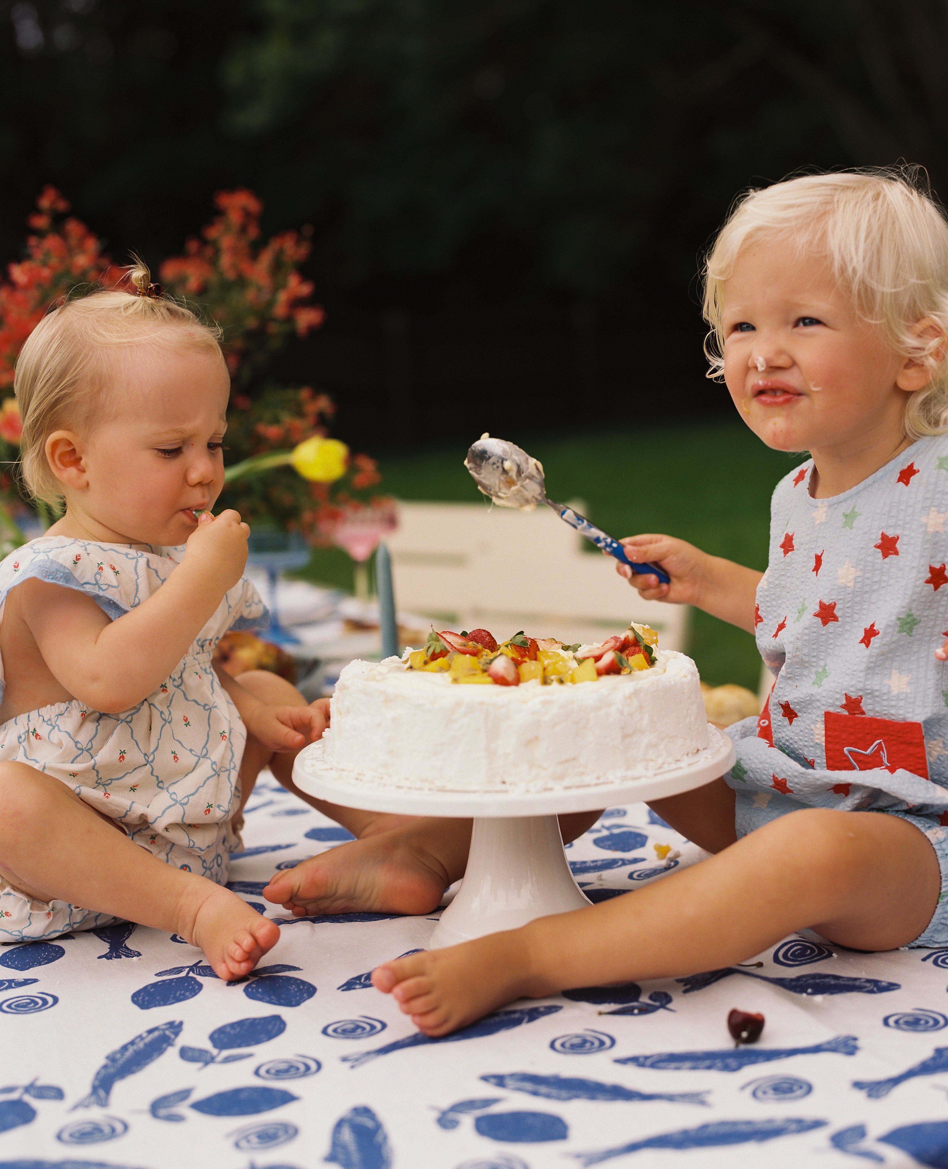 Two toddlers are seated on an outdoor table enjoying a cake adorned with fruit. One child is using a spoon, while the other samples with fingers. Both small ones are barefoot and dressed in Levi Rompers by JUNI JNR, surrounded by a summer setting with flowers and trees.