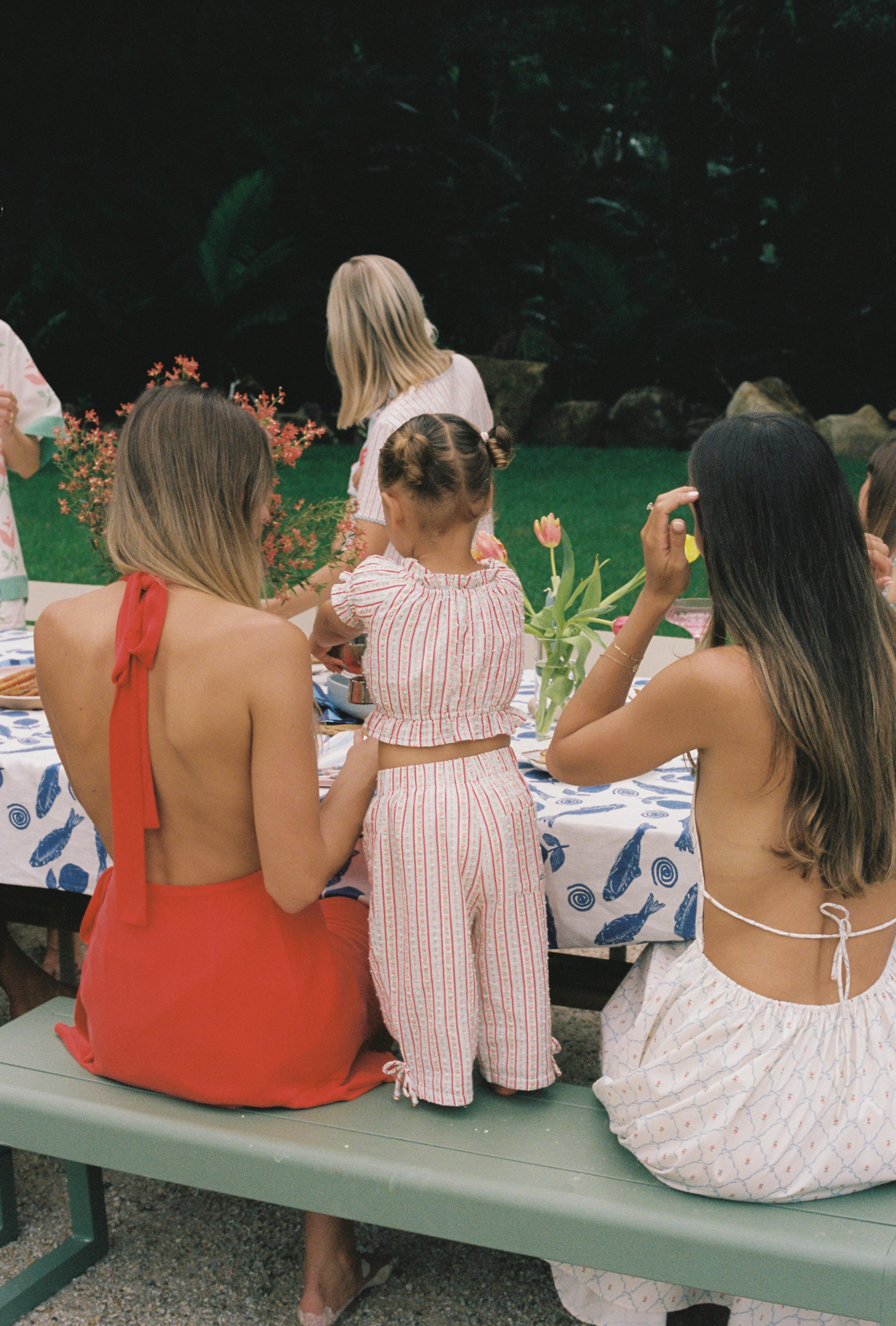 A group of people are seated at an outdoor table adorned with a blue patterned tablecloth made from textured cotton fabric. A child, wearing the Dahlia Top by JUNI JNR, stands on a bench between two adults, all dressed in summer outfits. Lush greenery is visible in the background, adding to the vibrant and relaxed atmosphere.