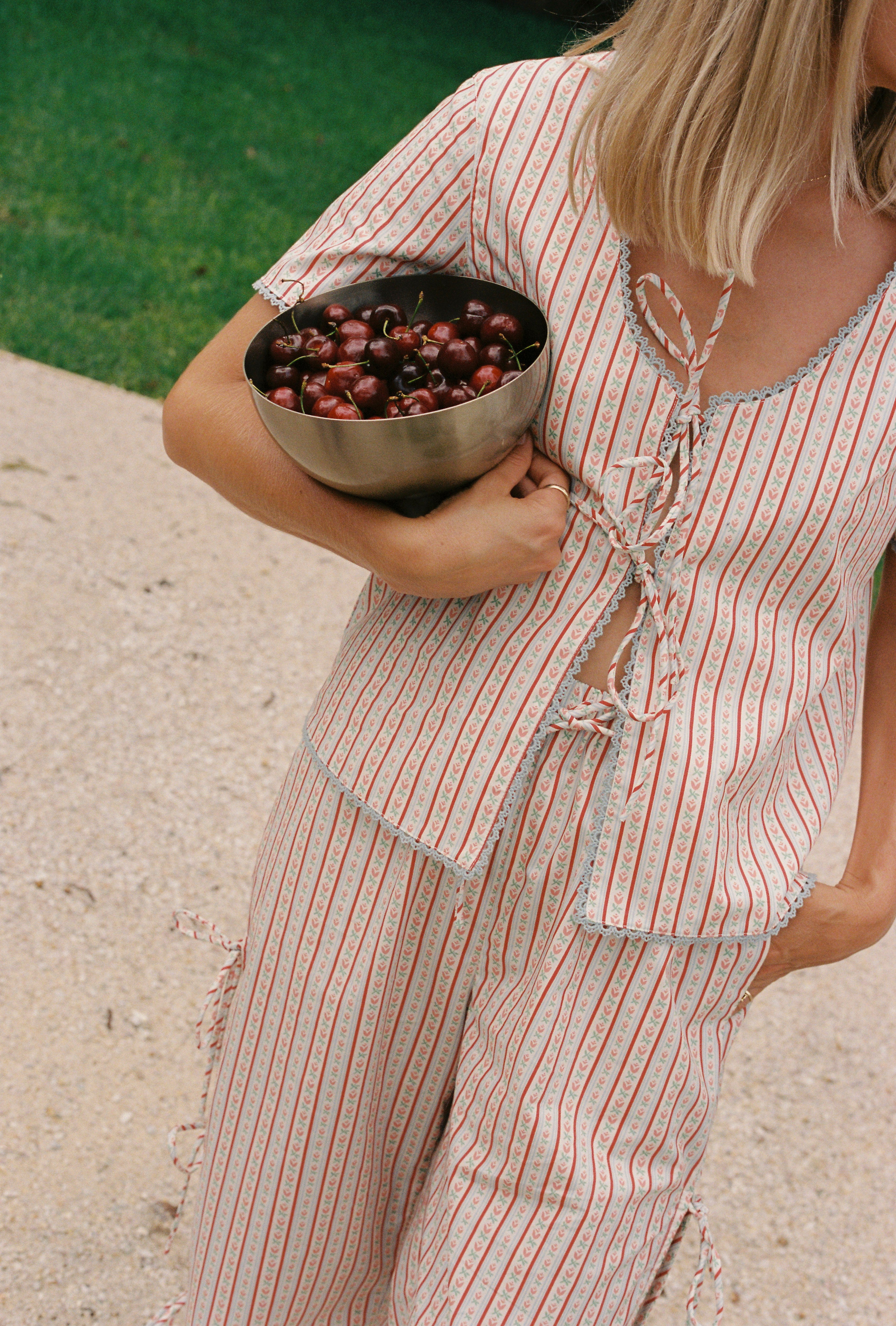 A person holding a metal bowl filled with cherries wears the Poppy Blouse by JUNI, a light-colored, striped outfit made of cotton poplin, featuring a tie-front top and matching pants. The background reveals a grassy area and a stone surface, enhancing the summery vibe.