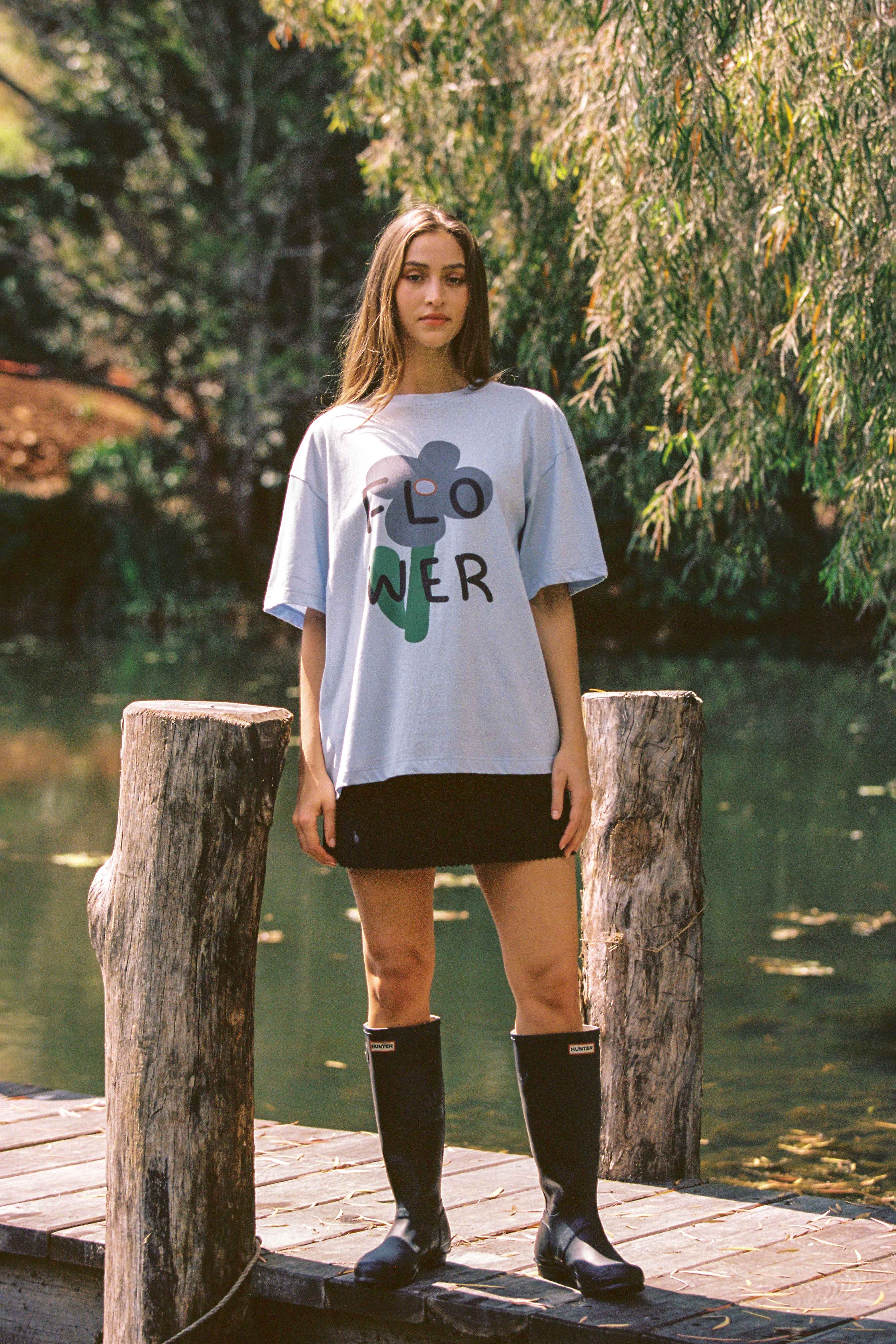 A woman stands on a wooden dock near a body of water surrounded by lush greenery. She wears the Bloom Tee in Light Blue by JUNI, featuring an ethically made, oversized design with "FLOWER" printed on it. She pairs the tee with a short black skirt and black knee-high rubber boots. She gazes directly at the camera, with trees in the background.