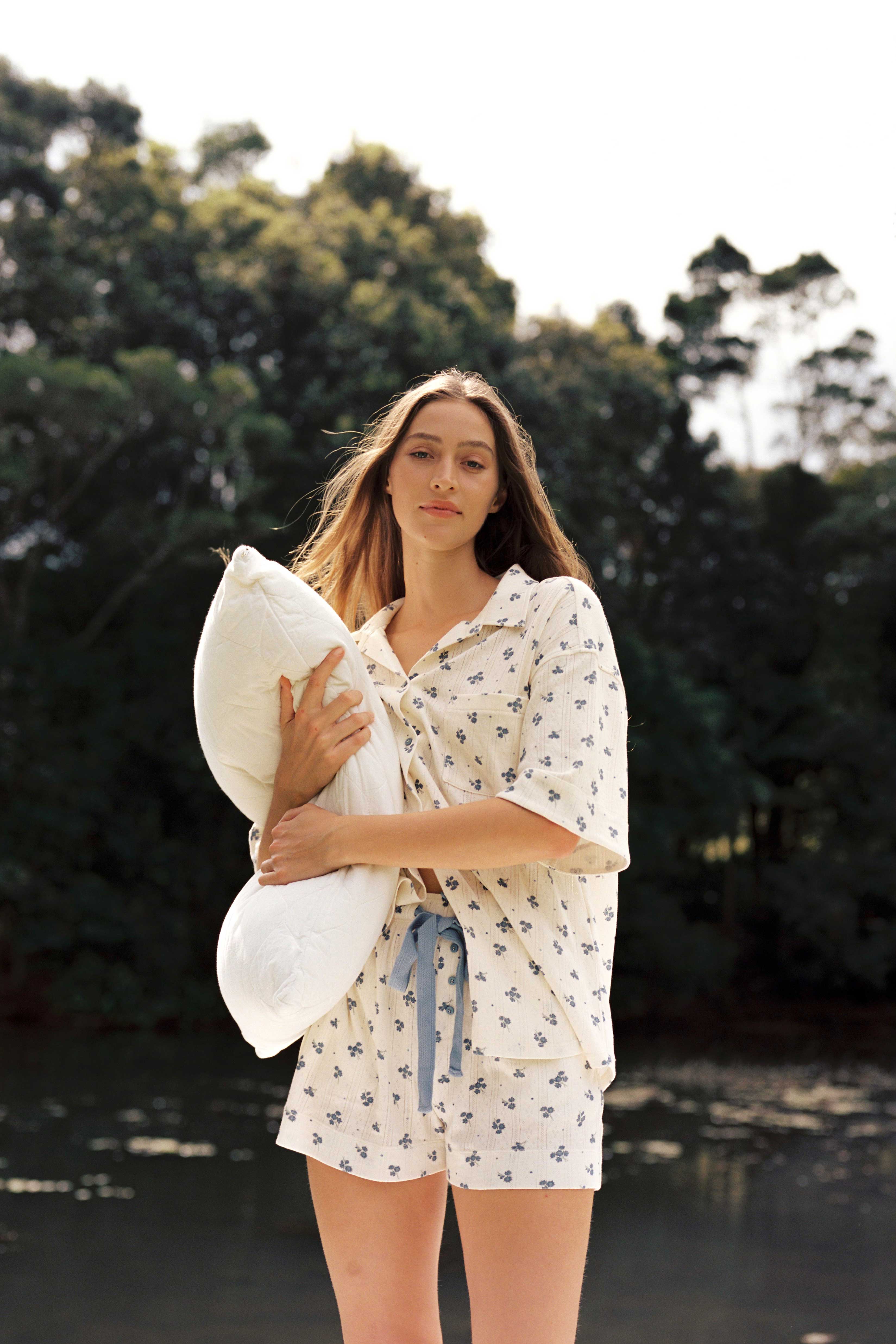 A woman wearing the Daze Pyjama Set from JUNI holds a pillow while standing outdoors near a body of water, with trees in the background. The light, floral pattern of her ethically made pajamas complements her long hair as she gazes forward with a calm expression.