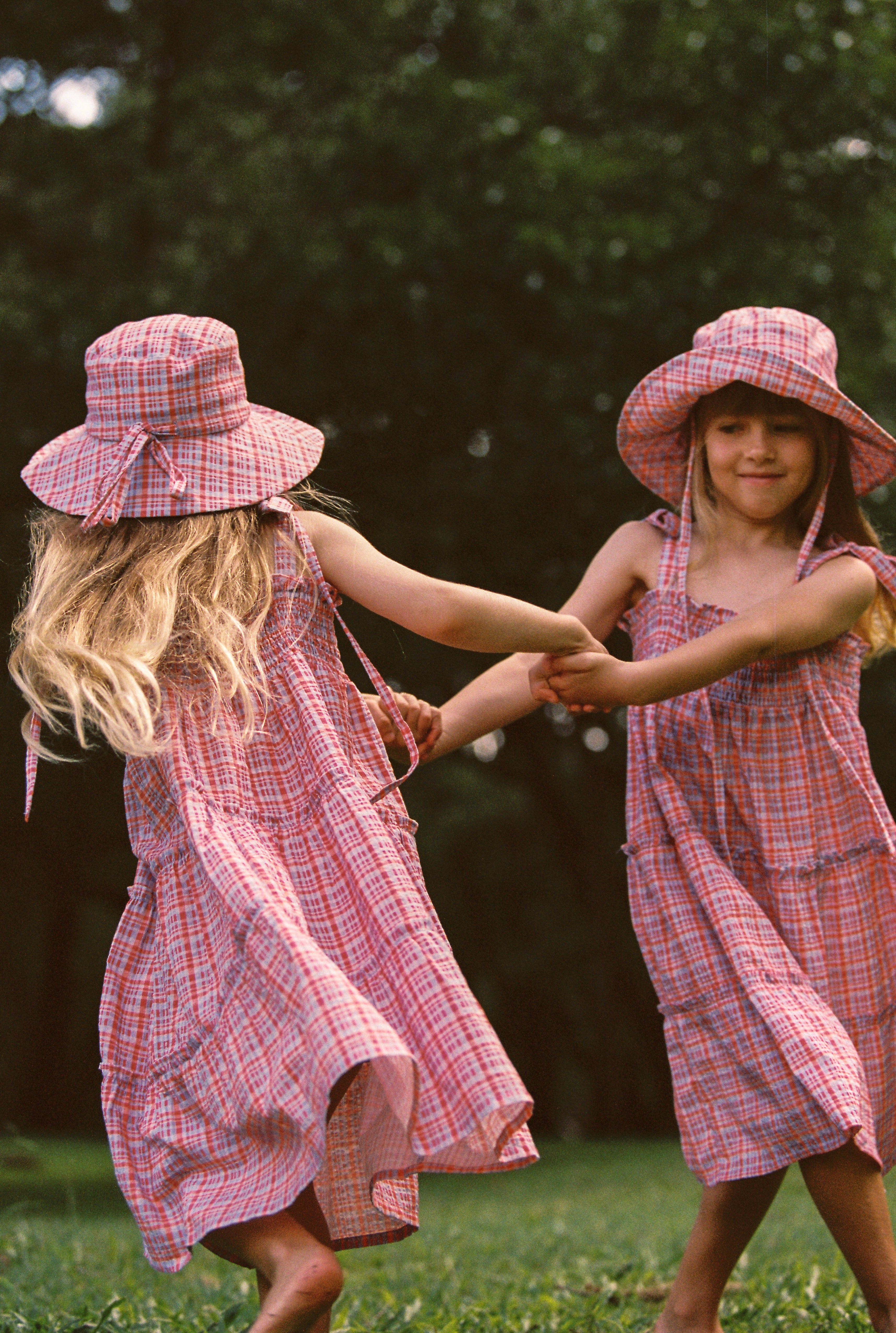 Two young girls dance joyfully on a grassy field in matching pink plaid dresses and JUNI JNR ~ Picnic Check Sunhats. Their long hair flows as they spin hand in hand, surrounded by a backdrop of trees.