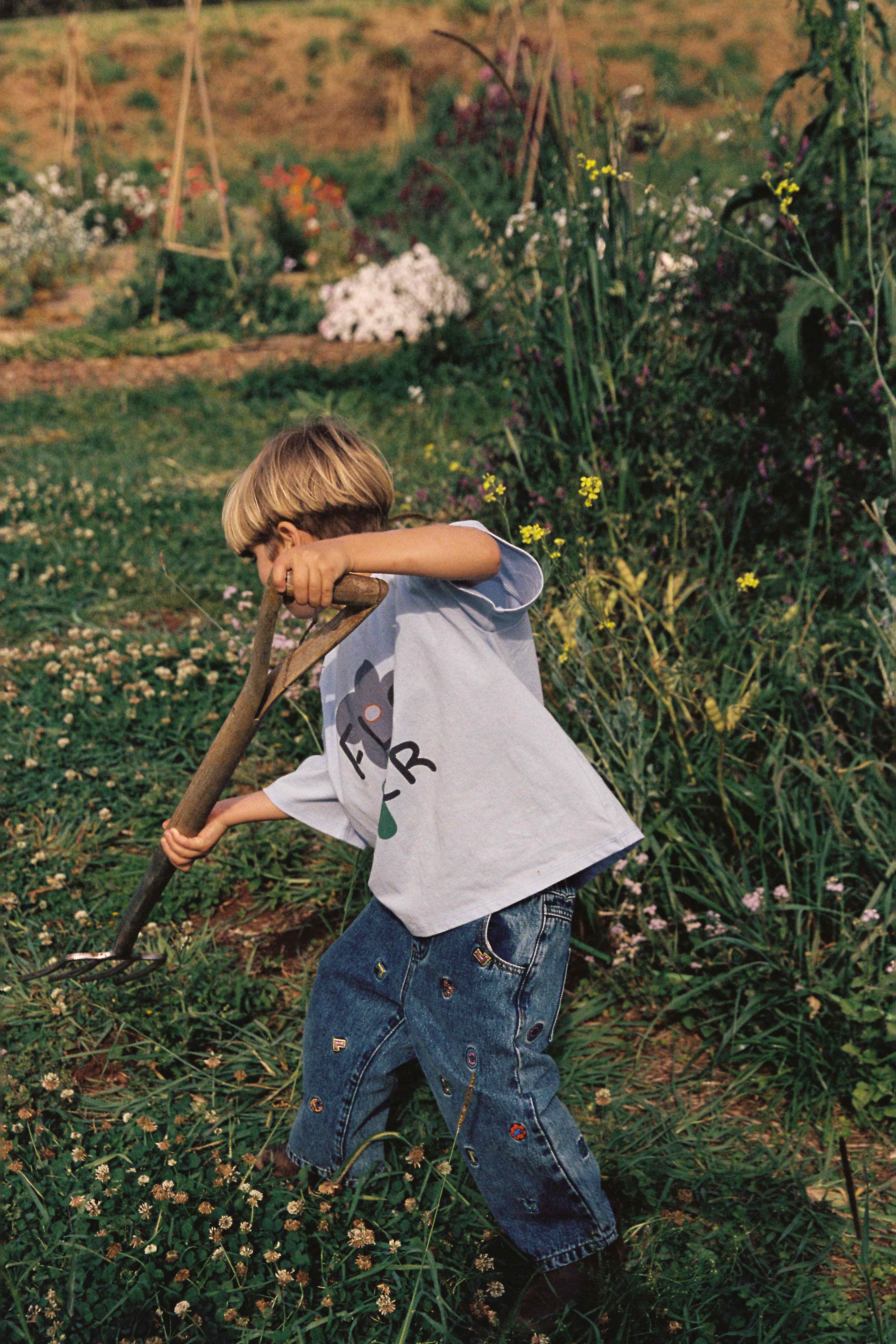 In a lush garden surrounded by vibrant greenery and colorful flowers, a young child wearing a light blue shirt and JUNI JUNIOR's Alphabet Denim Jean is energetically digging the ground with a long-handled tool. In the background, more plants and a raised garden bed can be seen.