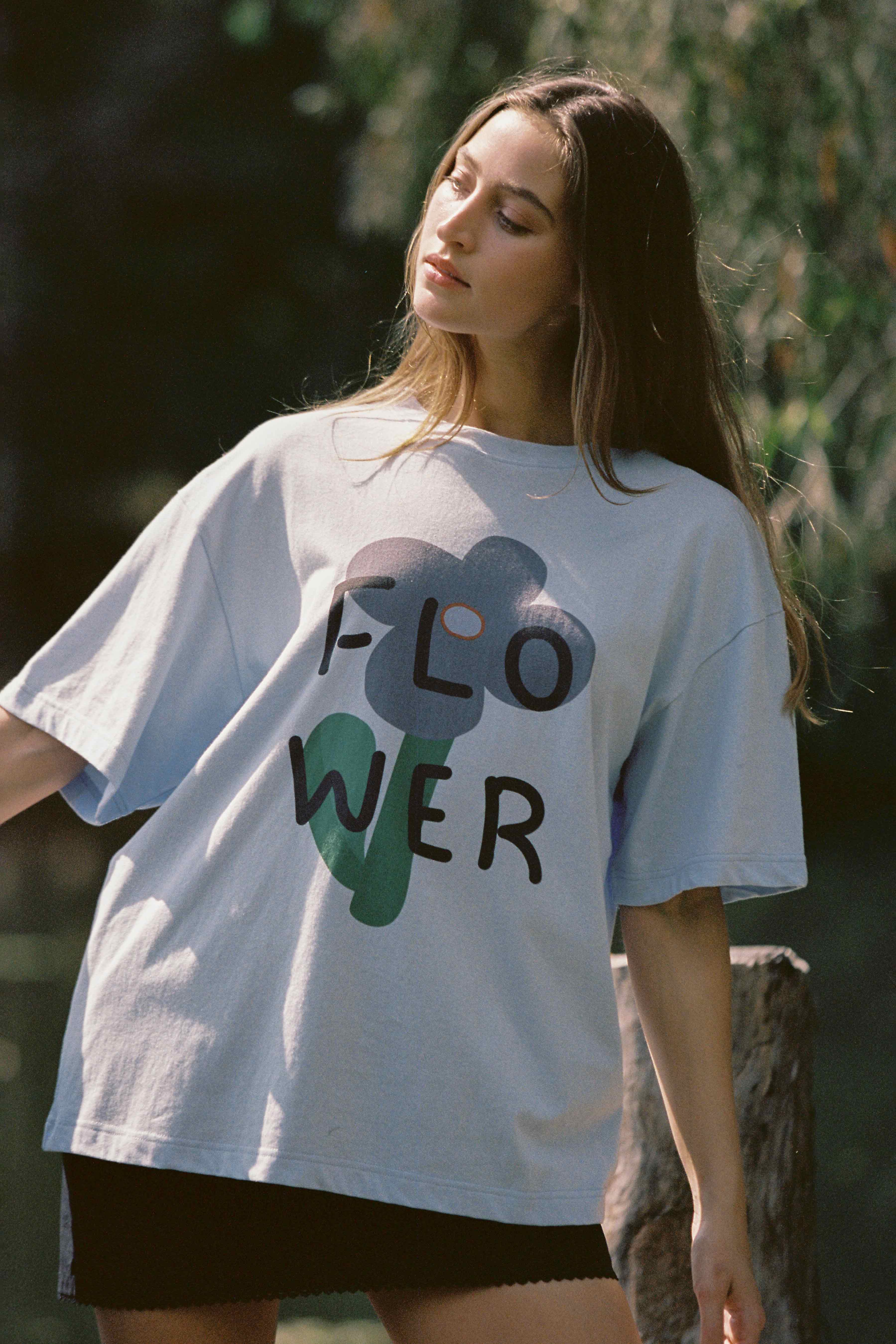 A young woman with long hair stands outdoors in a relaxed pose, wearing the Bloom Tee Light Blue from JUNI, an oversized fit T-shirt made from organic cotton. The word "FLOWER" is emblazoned in large, playful letters across the front. The background is blurred with greenery, suggesting a natural setting.