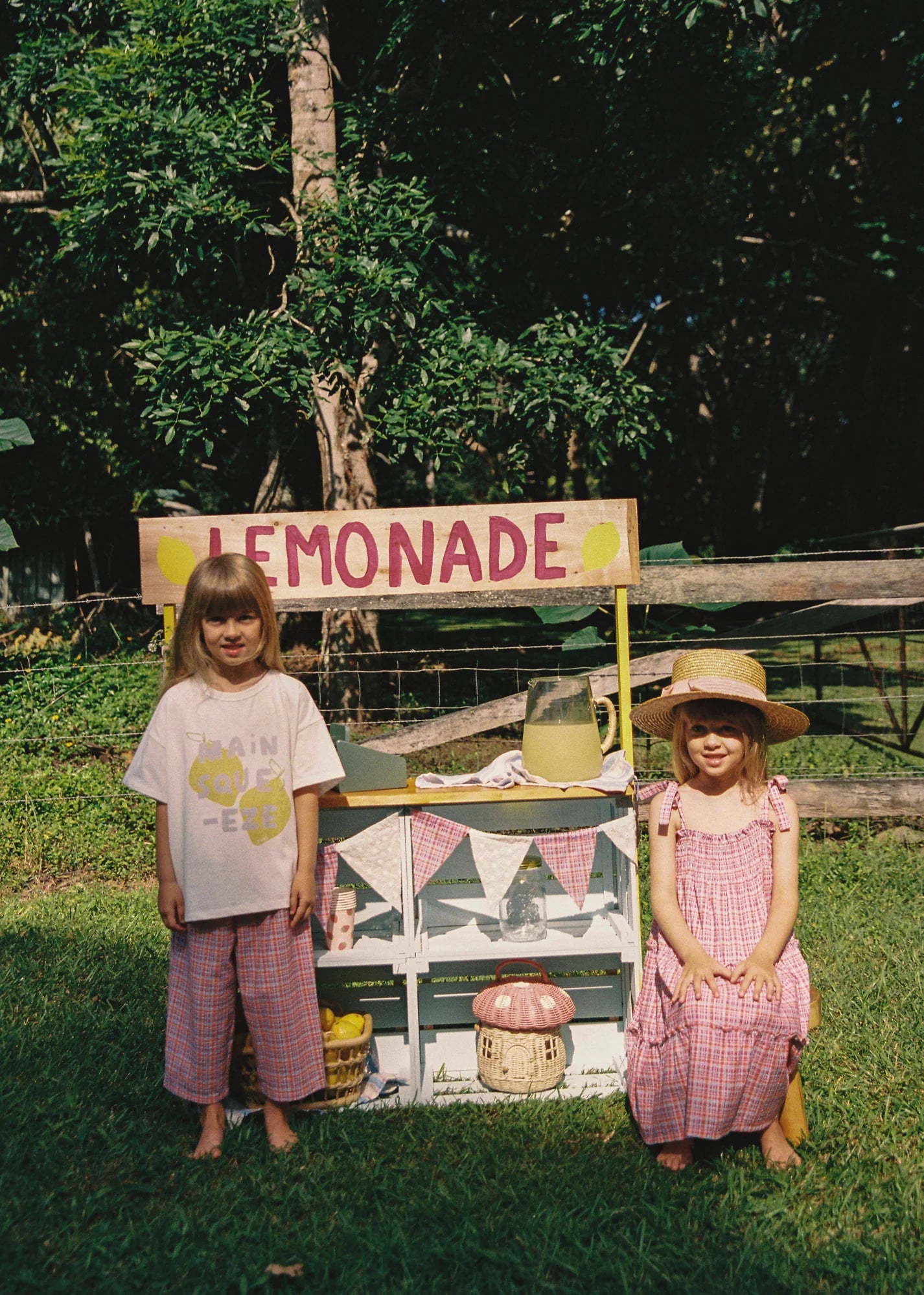 Two children are at a lemonade stand. One child wears JUNI JNR's Lenny Pant Picnic Check, while the other sits in a seersucker dress and straw hat. The stand has a "LEMONADE" sign and pitcher, with trees and a fence in the background.