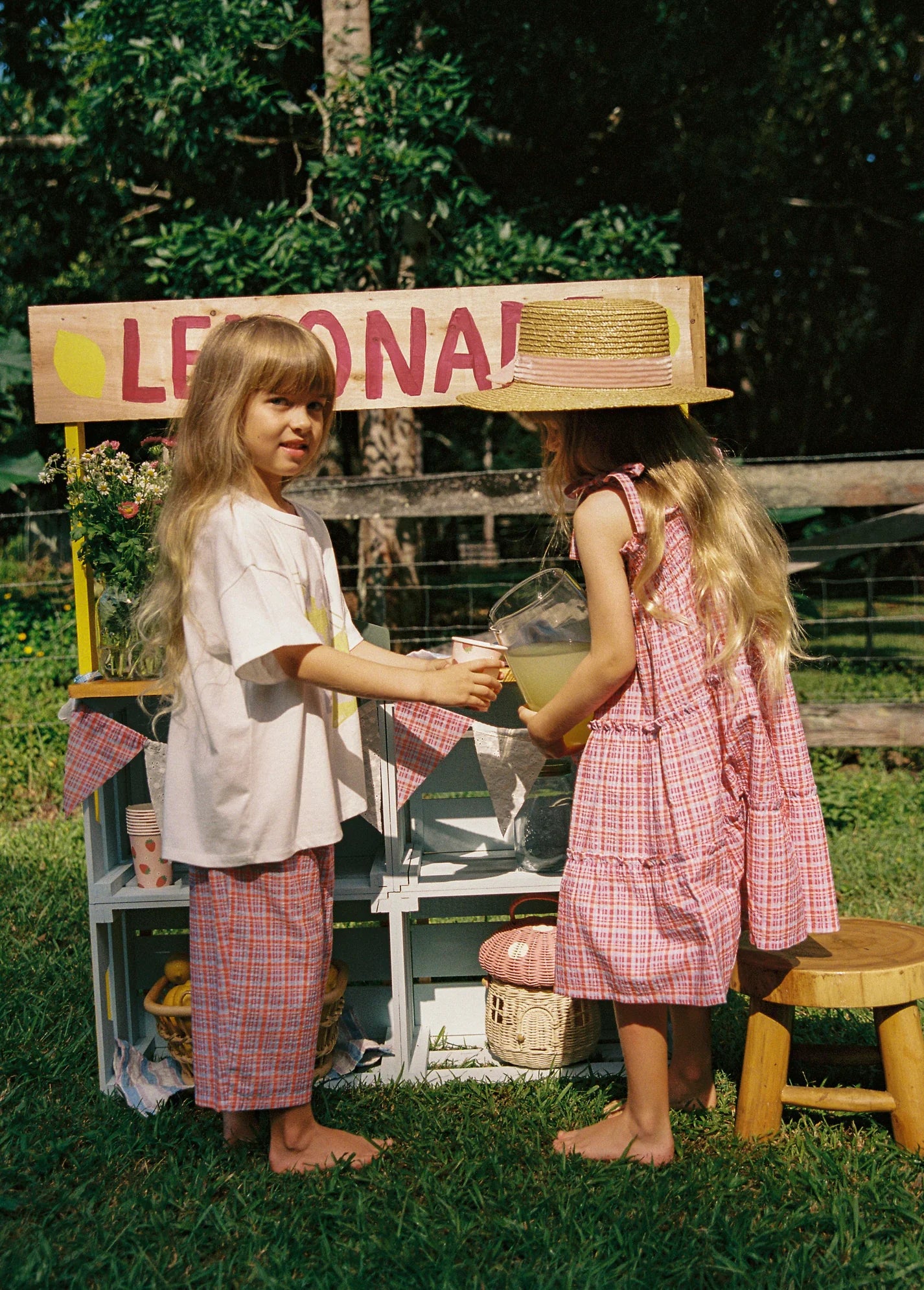 Two young children with long hair are at a lemonade stand. One, in a pink plaid dress and straw hat, pours lemonade for the other wearing JUNI JNR's Lenny Pant Picnic Check with an elastic waistband. The stand is labeled "Lemonade" and set outdoors amidst lush greenery.