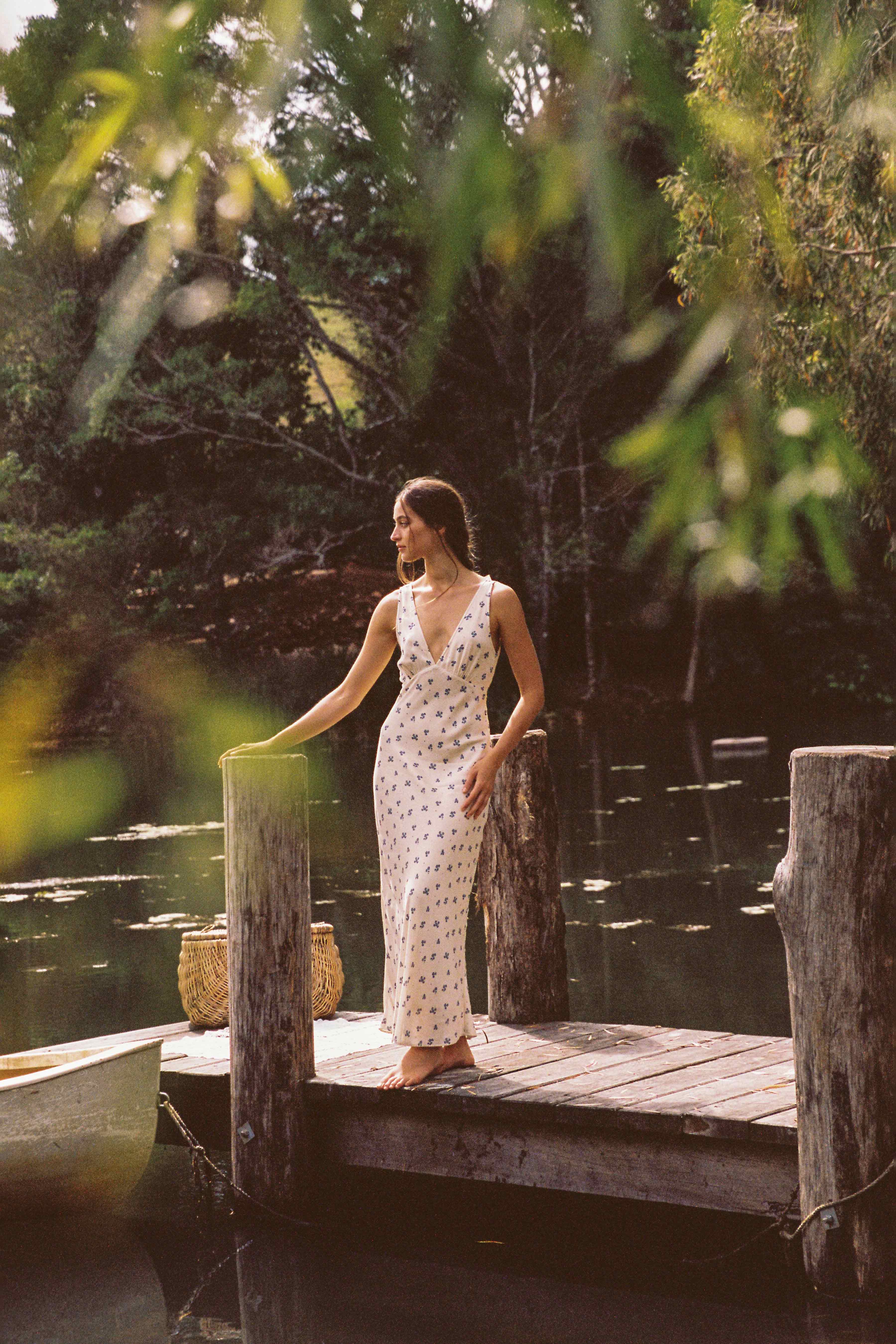 Wearing the JUNI Isabella Midi Dress Ditsy Floral, a woman stands barefoot on a wooden dock, touching a post. The sleeveless dress features a v-neckline and has an elegant ditsy floral pattern. She gazes into the distance at the serene lake surrounded by lush greenery, with a wicker basket nearby.