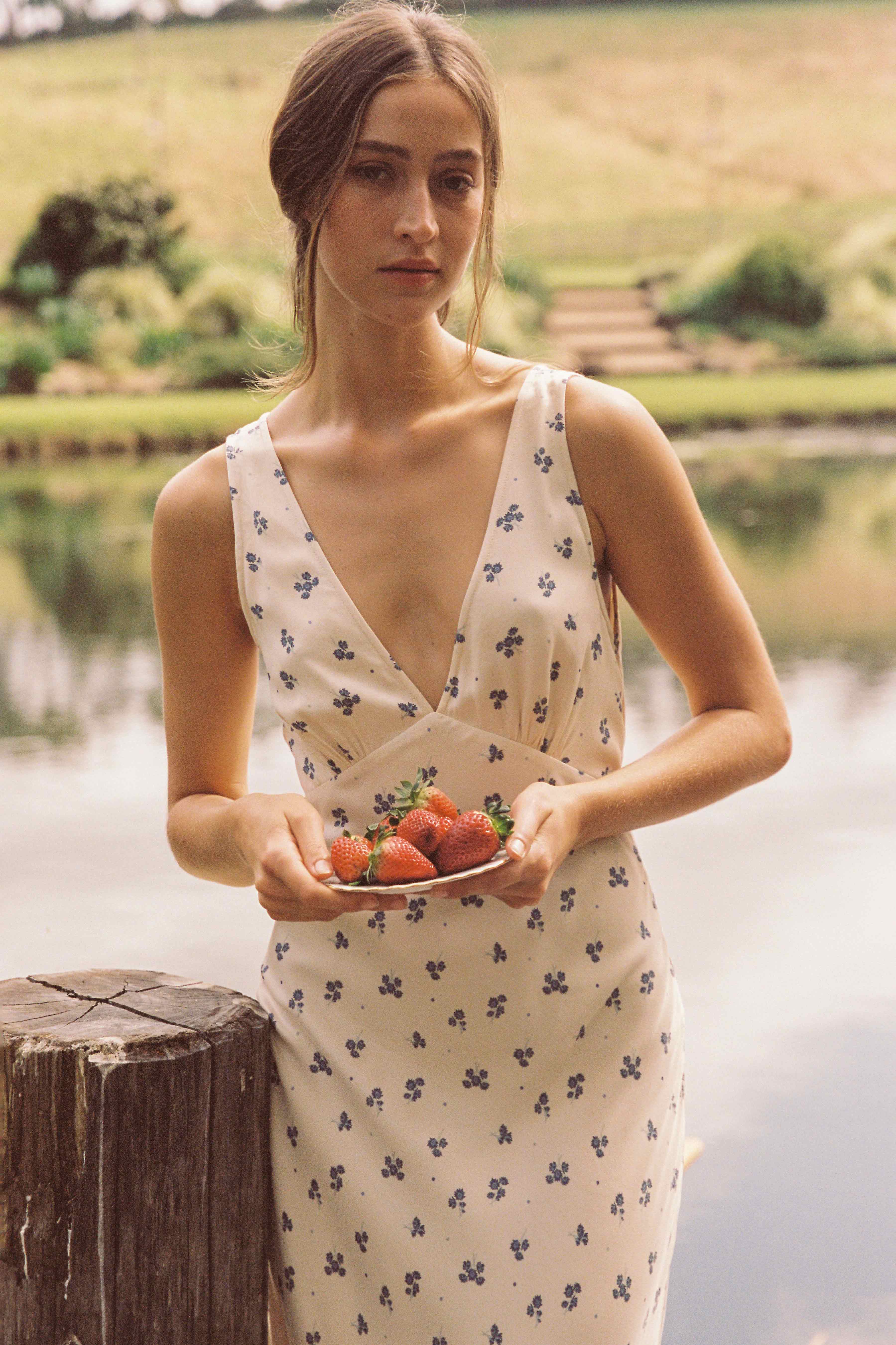 A person with long hair stands outdoors by a wooden post holding a plate of strawberries. They are wearing the ethically made Isabella Midi Dress Ditsy Floral by JUNI, which features a sleeveless design with a v neckline and floral pattern. In the background, there is a grassy landscape and a body of water, creating a serene and natural scene.