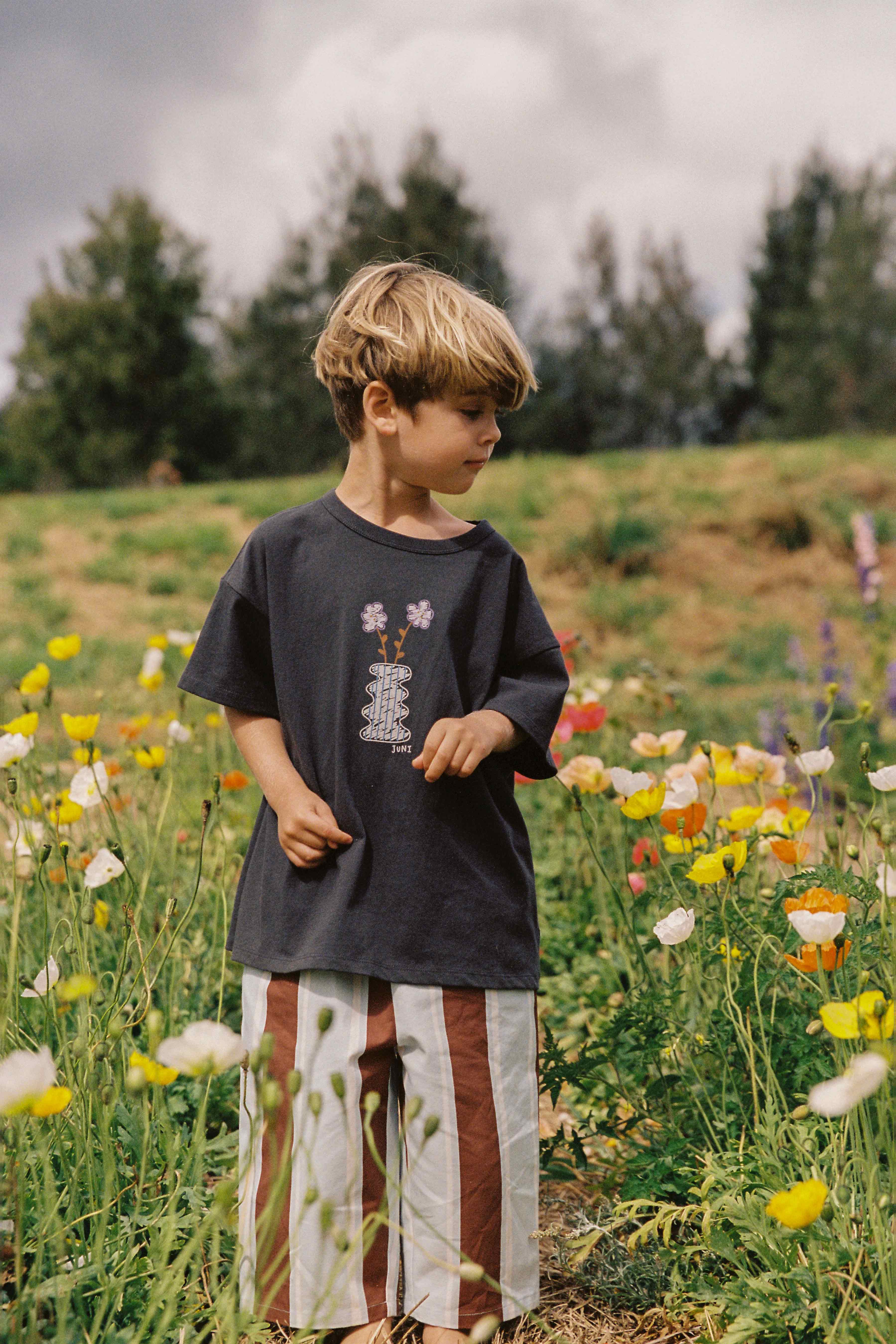 A young child with short blonde hair stands in a vibrant flower field, dressed in the Lenny Pant Watson Stripe from JUNI JUNIOR. The child is wearing a dark shirt with a floral design, and the striped wide-leg pants feature an elastic waistband. Behind them are tall green trees and a cloudy sky. The child's gaze is slightly downward as they enjoy their natural surroundings.