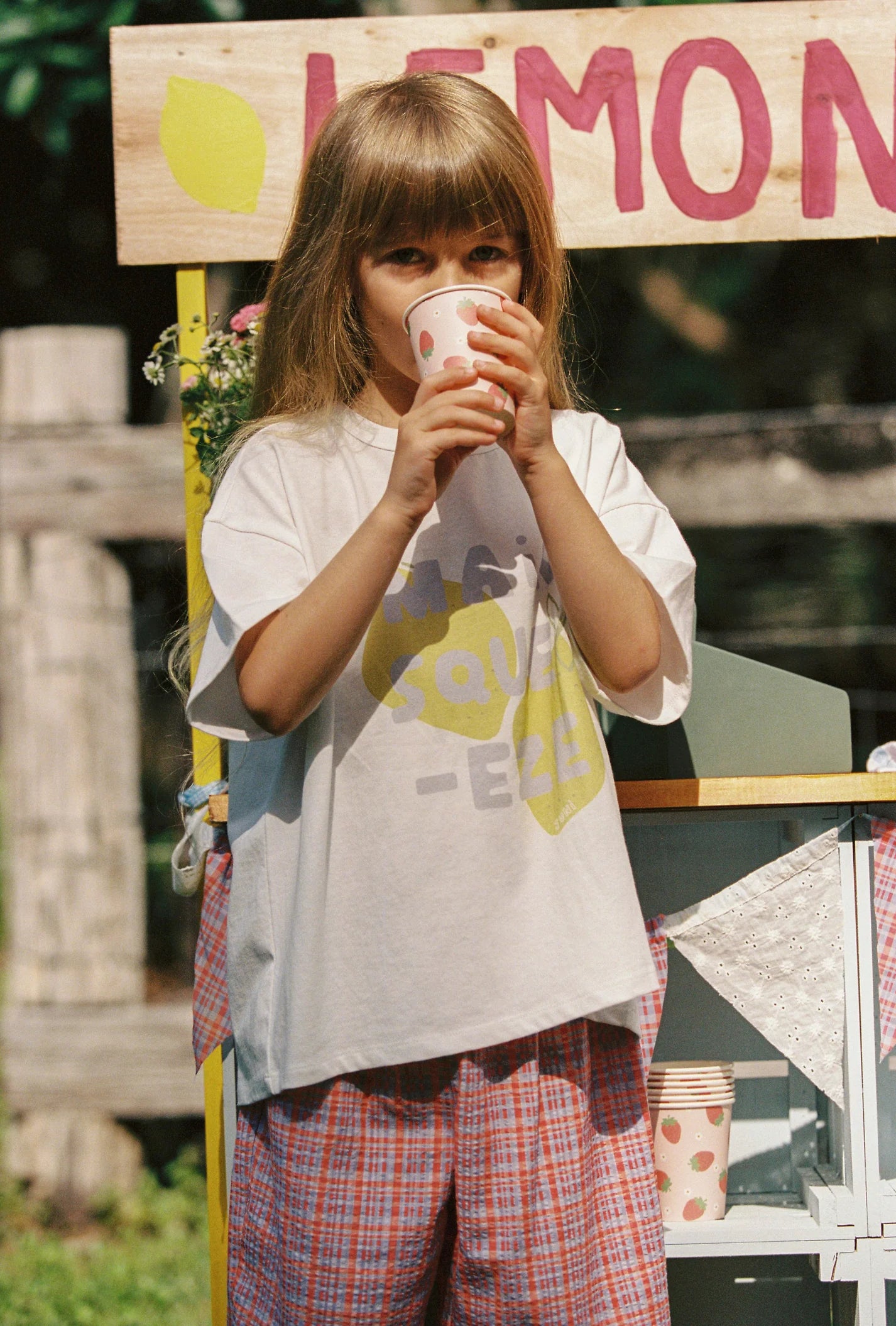 A young girl with long hair sips from a cup at a lemonade stand, wearing JUNI JNR's Main Squeeze Tee made of GOTS certified organic cotton and red plaid pants. The stand has handwritten signs and cups, with a wooden fence in the background.