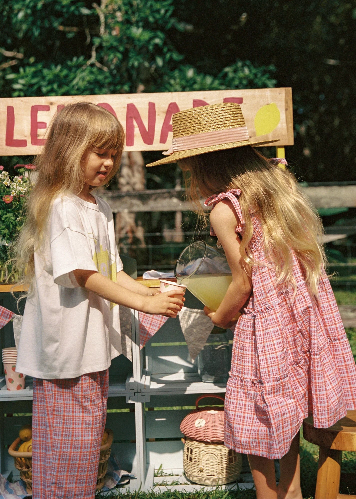 Two young girls run a lemonade stand outside. One, in a straw hat and plaid dress, pours lemonade into a cup held by the other, wearing JUNI JNR's ethically made Main Squeeze Tee and plaid pants. A wooden sign above reads "LEMONADE.