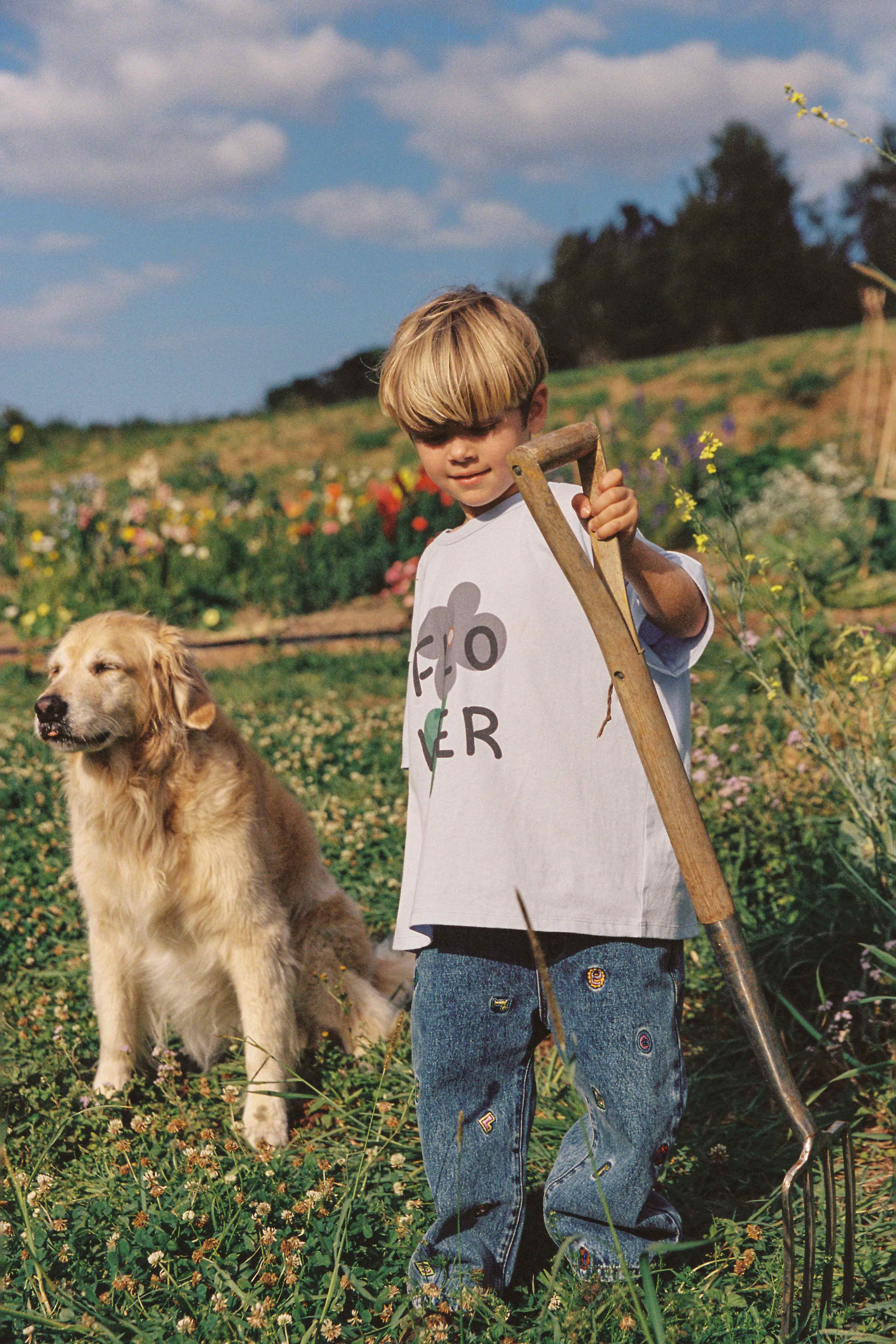 A young child stands in a grassy field holding a garden fork, with a golden retriever sitting nearby. The child wears a white t-shirt with the text "GROW LOVER" and JUNI JUNIOR's Alphabet Denim Jean, decorated with small patches and intricate embroidery. The background features a lush garden under a partly cloudy sky.