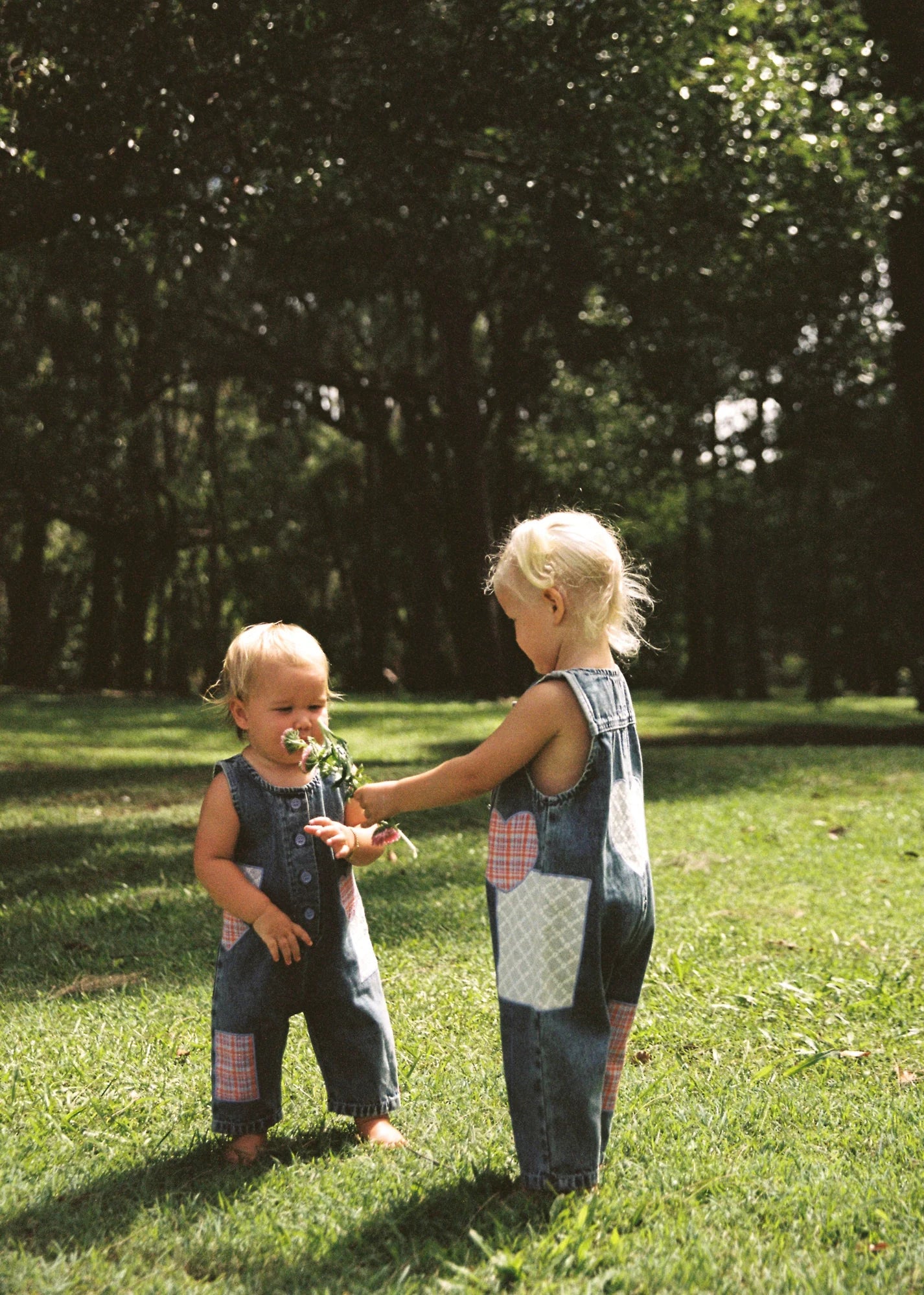 In a sunlit park, two blonde toddlers stand on grass wearing JUNI JNR ~ Charlie Overalls. One hands the other a bunch of flowers, surrounded by trees and greenery, all while sporting these ethically made denim overalls with vibrant patchwork designs.