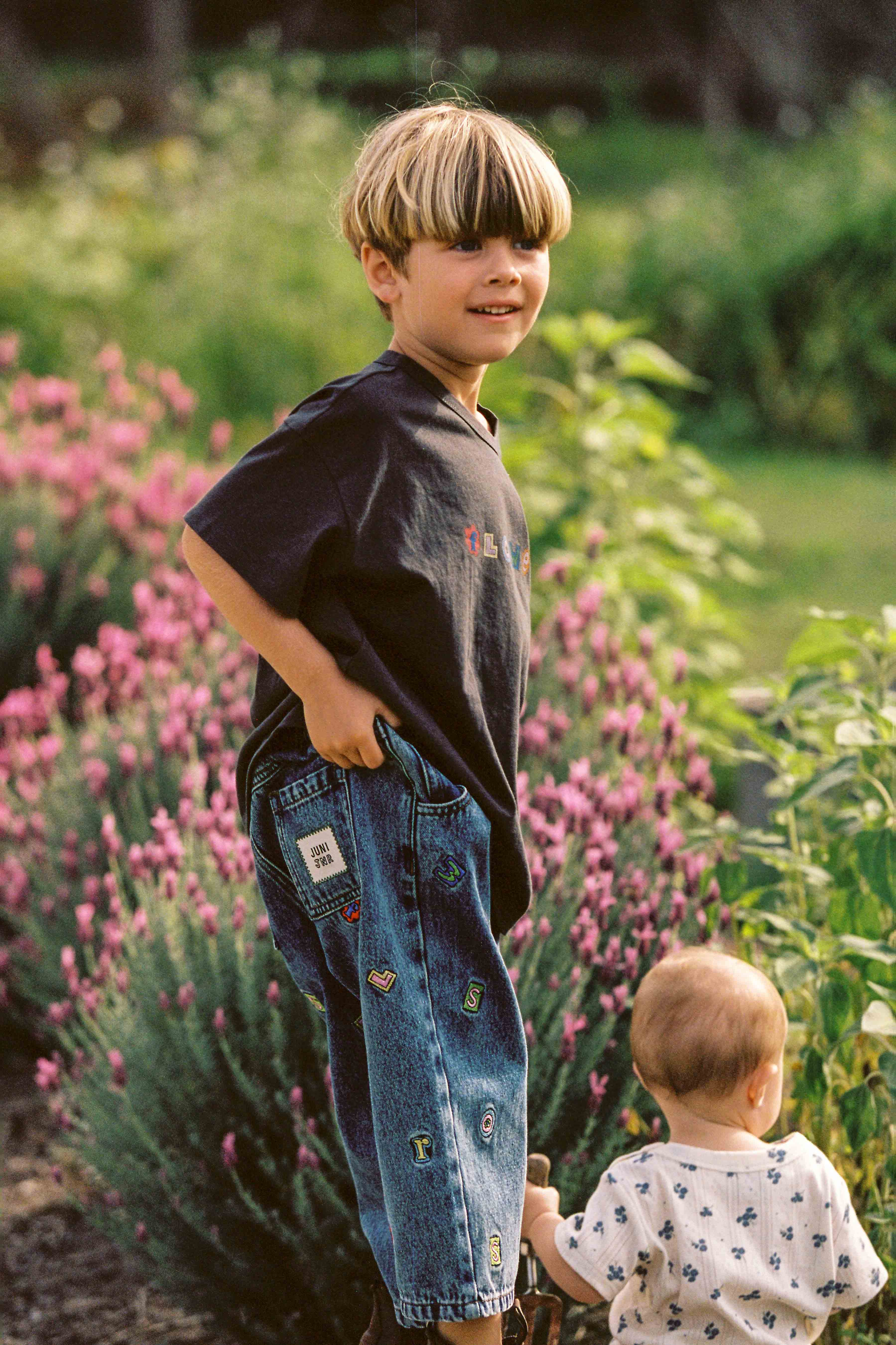 A young boy with a bowl haircut, dressed in a dark t-shirt and JUNI JUNIOR's Alphabet Denim Jeans featuring patches and an elastic waistband, stands outdoors with one hand in his pocket, smiling. Beside him is a baby in a white shirt adorned with embroidery, touching a green plant. Pink flowers and greenery create a colorful backdrop.