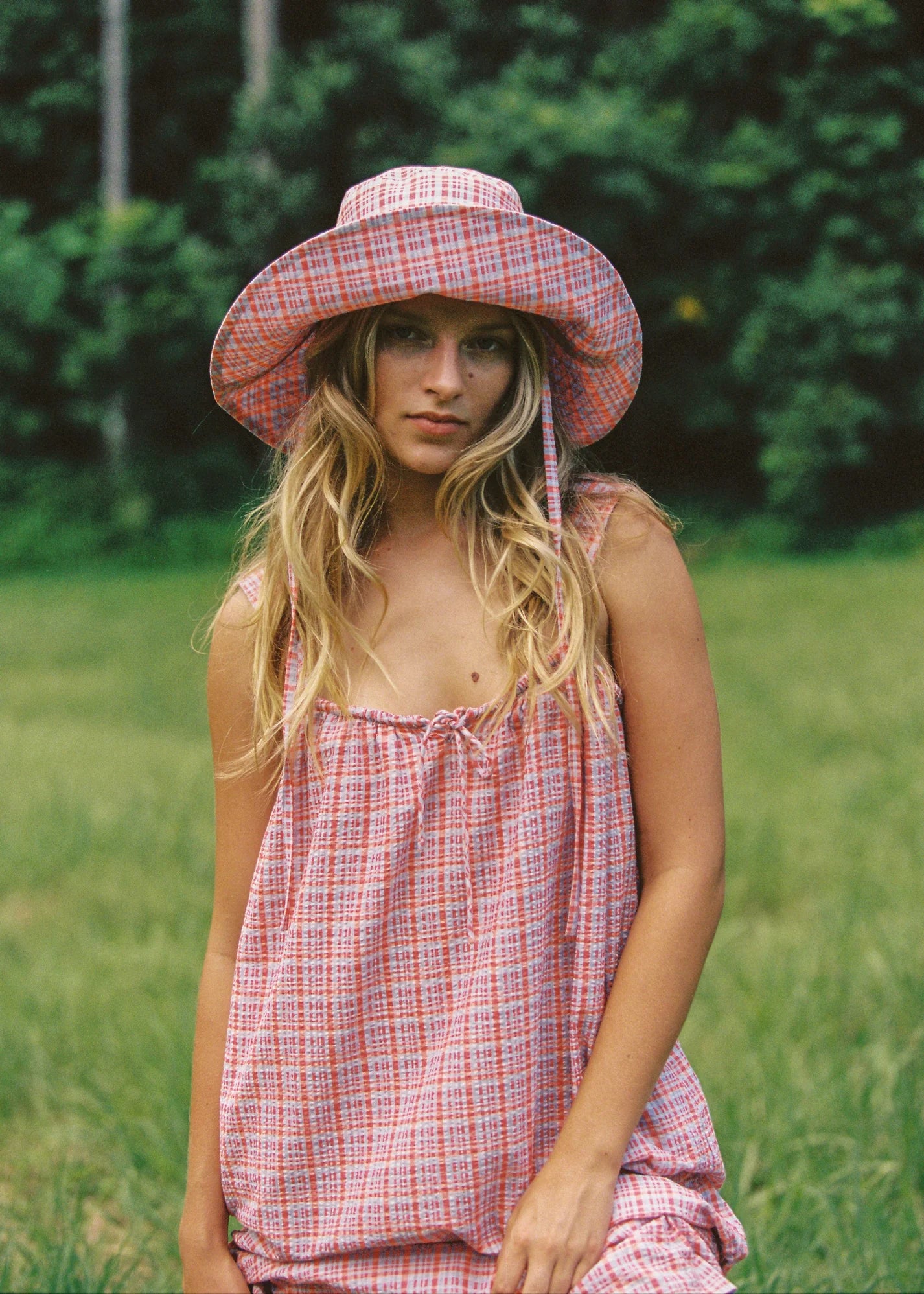 A person with long hair is in a grassy field, wearing the JUNI ~ Picnic Check Sunhat and a sleeveless red and white checkered dress featuring an exclusive print. The background is lush with green foliage.