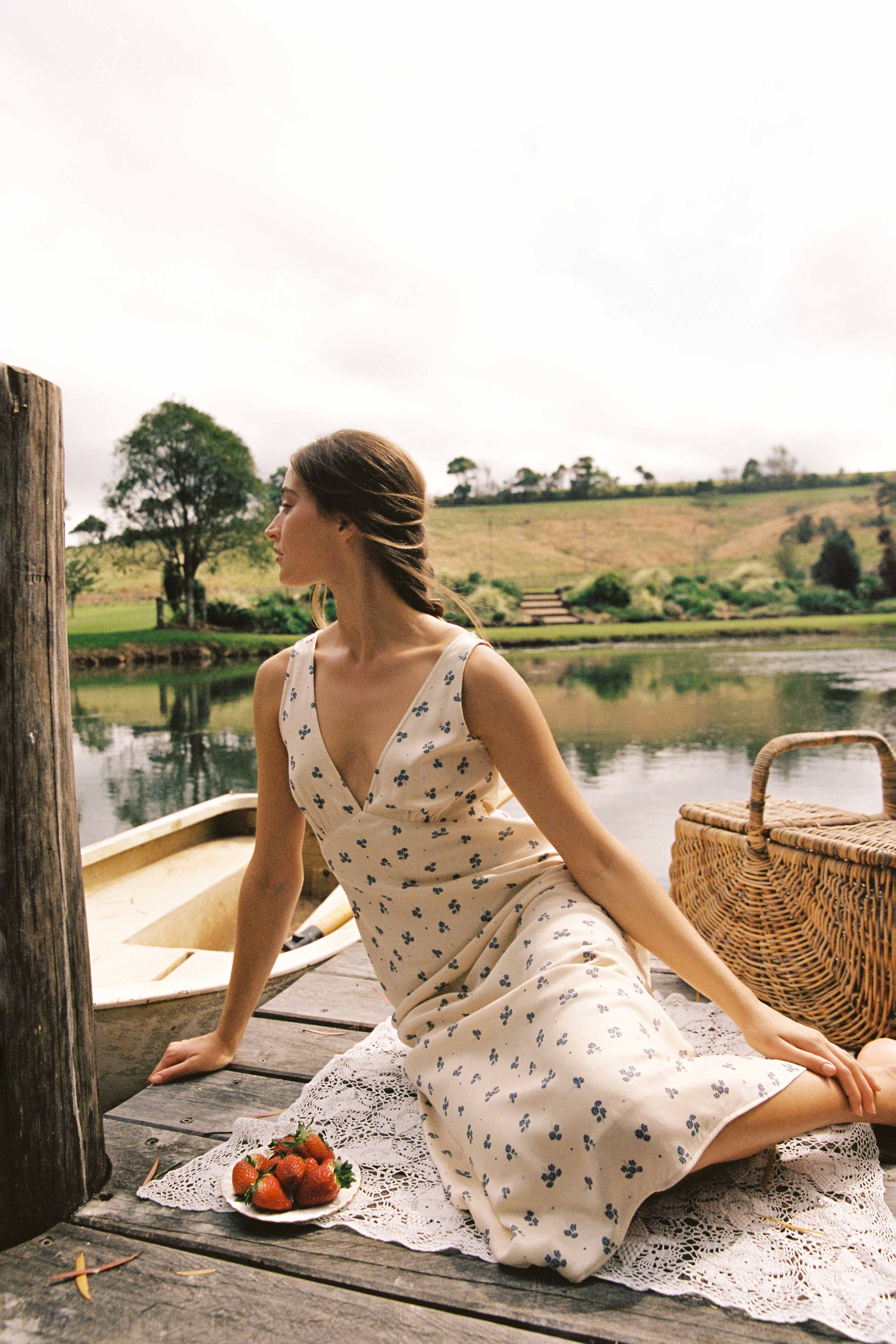 A woman in the ethically made Isabella Midi Dress Ditsy Floral by JUNI sits on a wooden dock by a tranquil lake. Beside her are a wicker basket and a plate of strawberries. She gazes to the side as gentle greenery and hills surround the serene water.