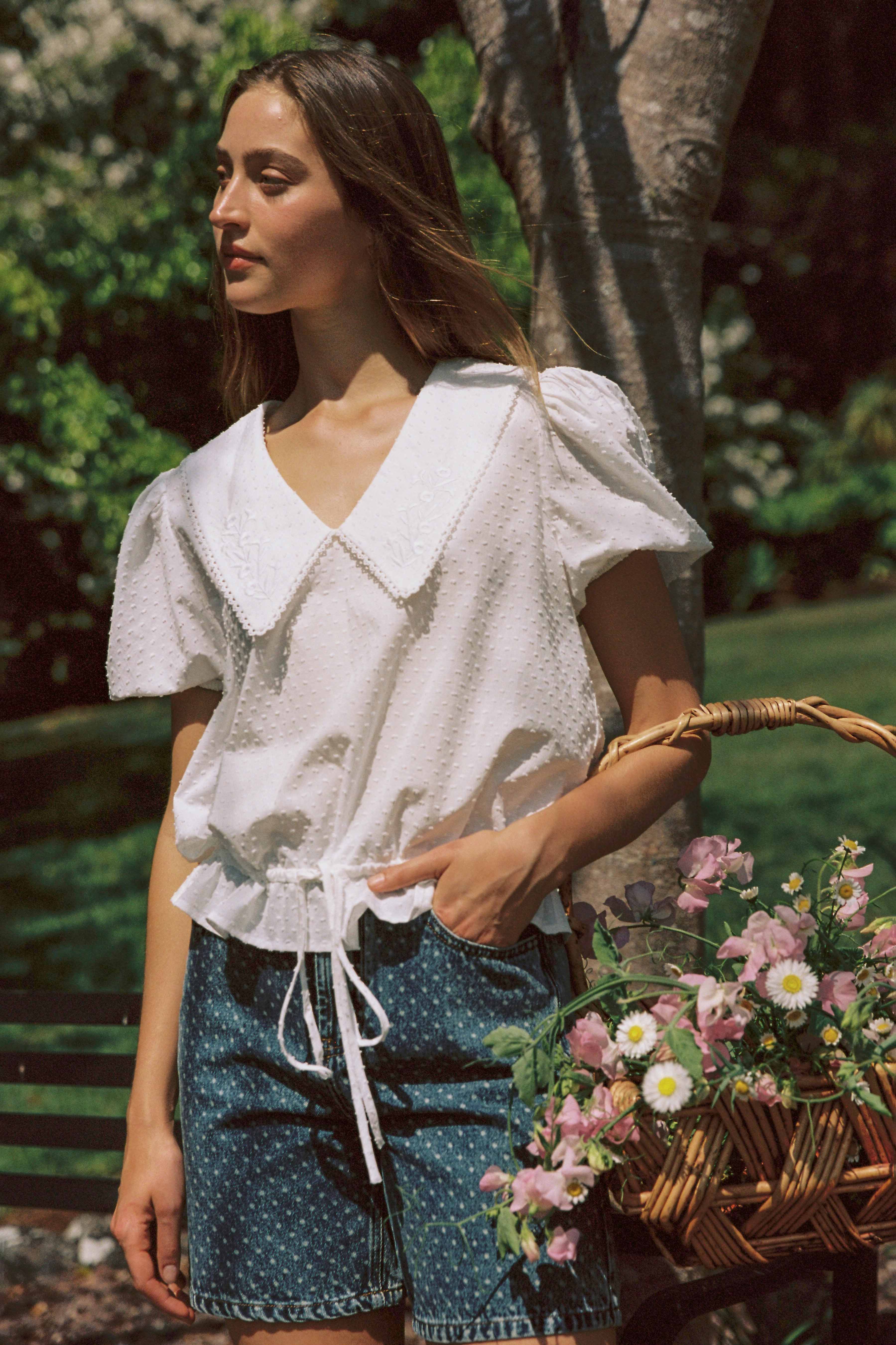 A person with long hair stands outdoors, donning a white short-sleeved blouse with a wide collar and wearing the "Bud Denim Short Spot" from JUNI, which features functional pockets. They are holding a wicker basket filled with pink and white flowers, with a tree and greenery visible in the background.