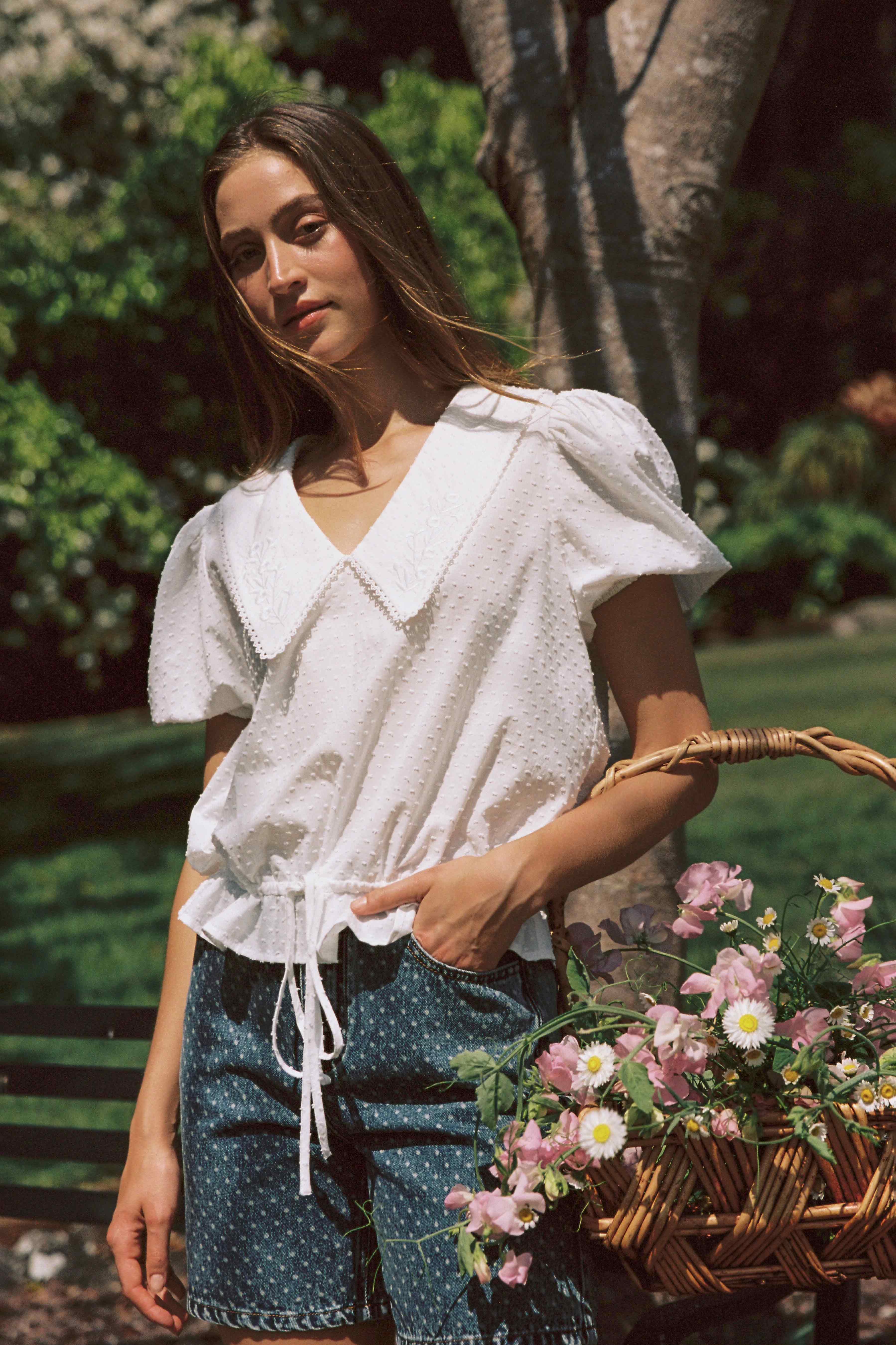 A woman with long brown hair stands outdoors near a tree, wearing a white blouse with puffed sleeves and JUNI's Bud Denim Short Spot. She holds a wicker basket filled with pink and white flowers. Sunlight filters through the greenery, casting soft shadows.